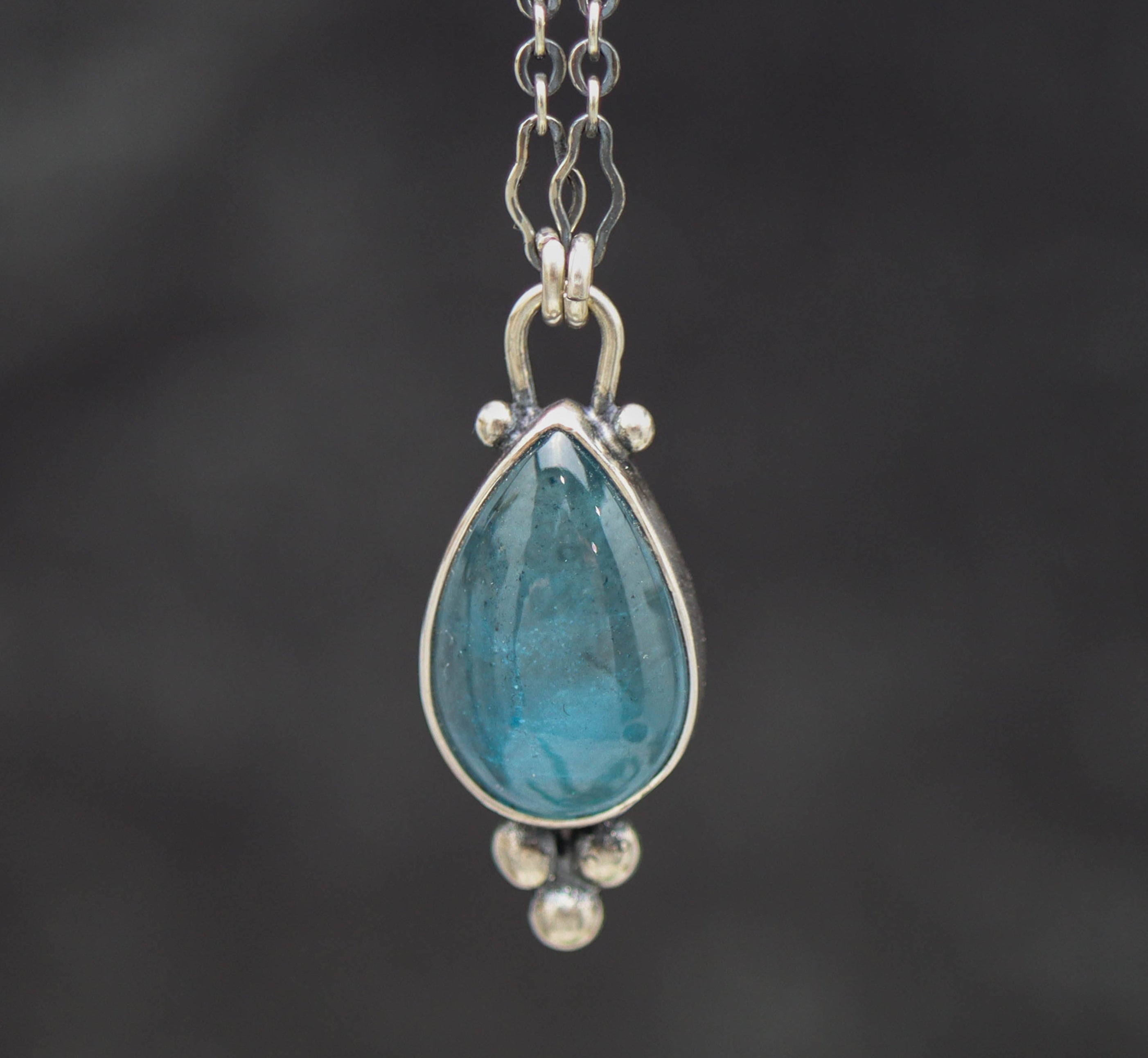 Teardrop-shaped blue topaz gemstone pendant on a chain against a dark background