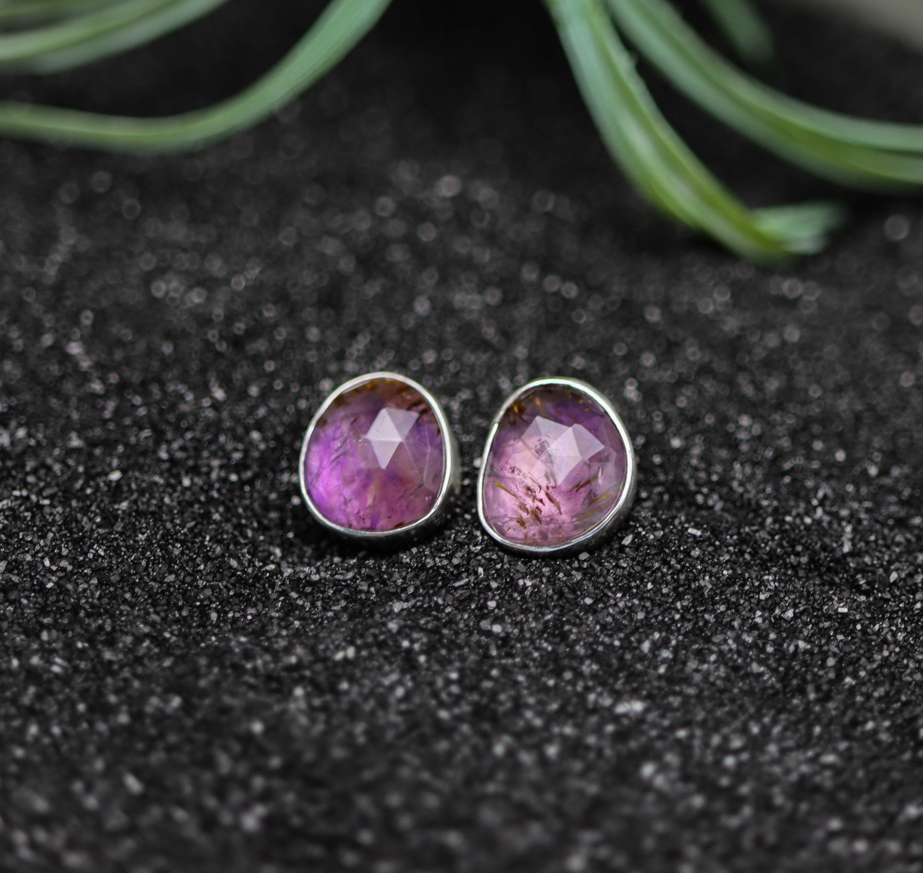 Purple gemstone stud earrings on a dark textured surface with green leaves in the background