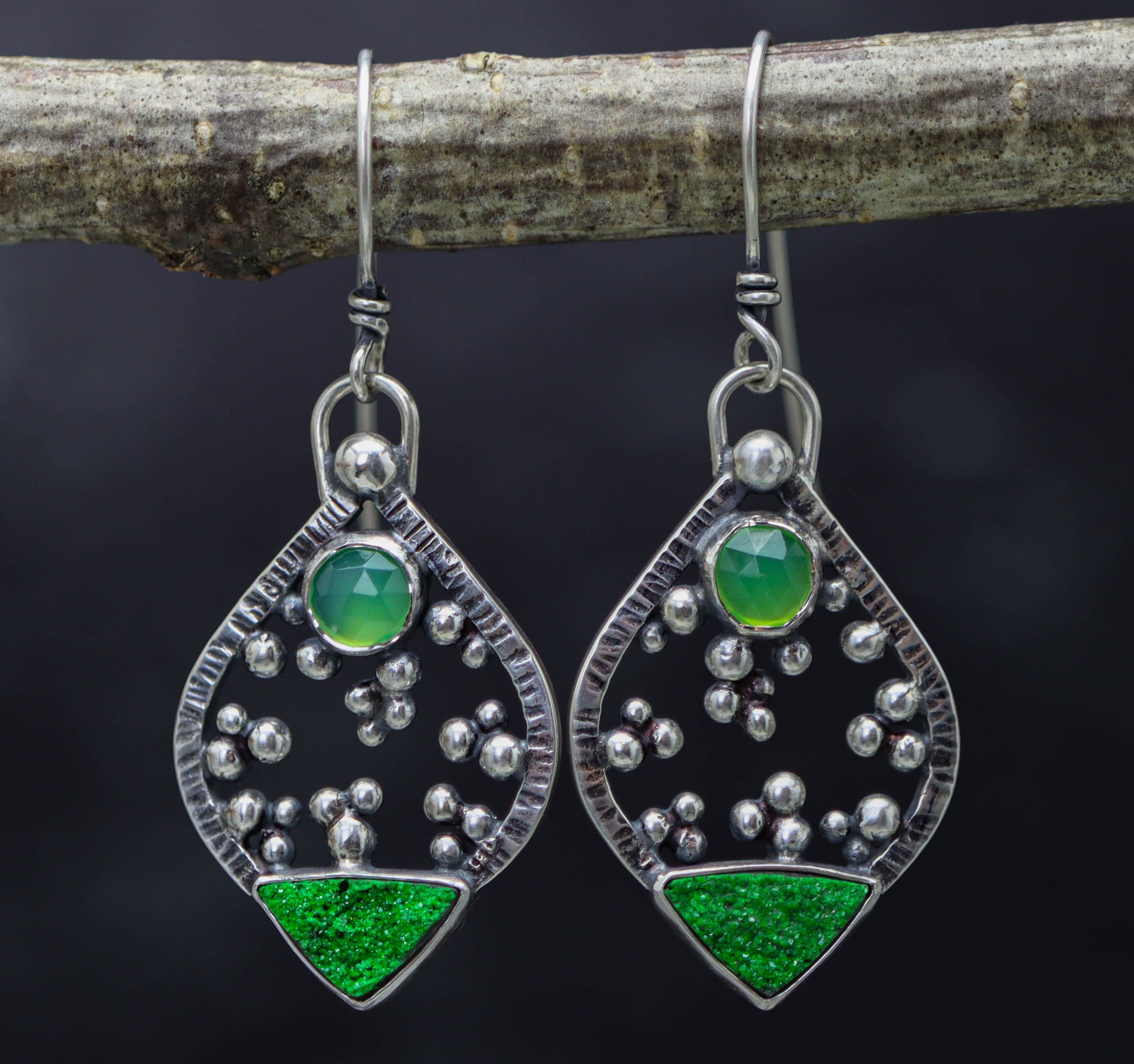 Pair of silver earrings with green gemstones hanging on a branch against a dark background