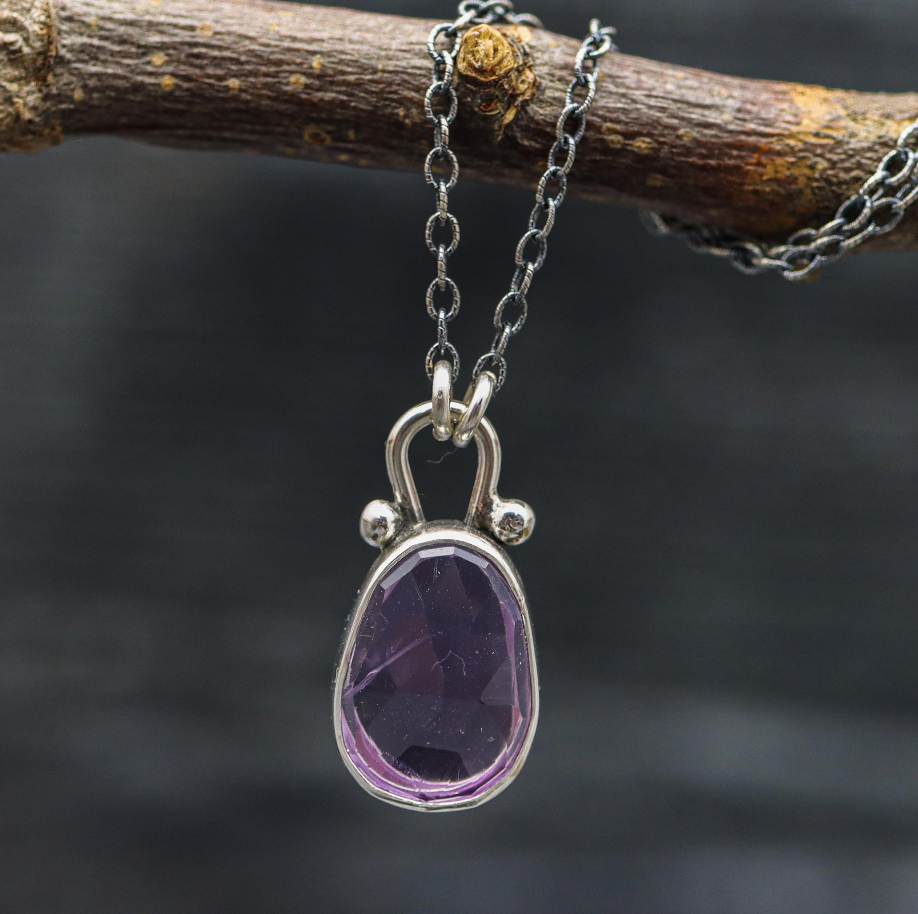 A sterling silver pendant necklace with a faceted rose cut amethyst centerpiece, displayed on a branch against a dark background.