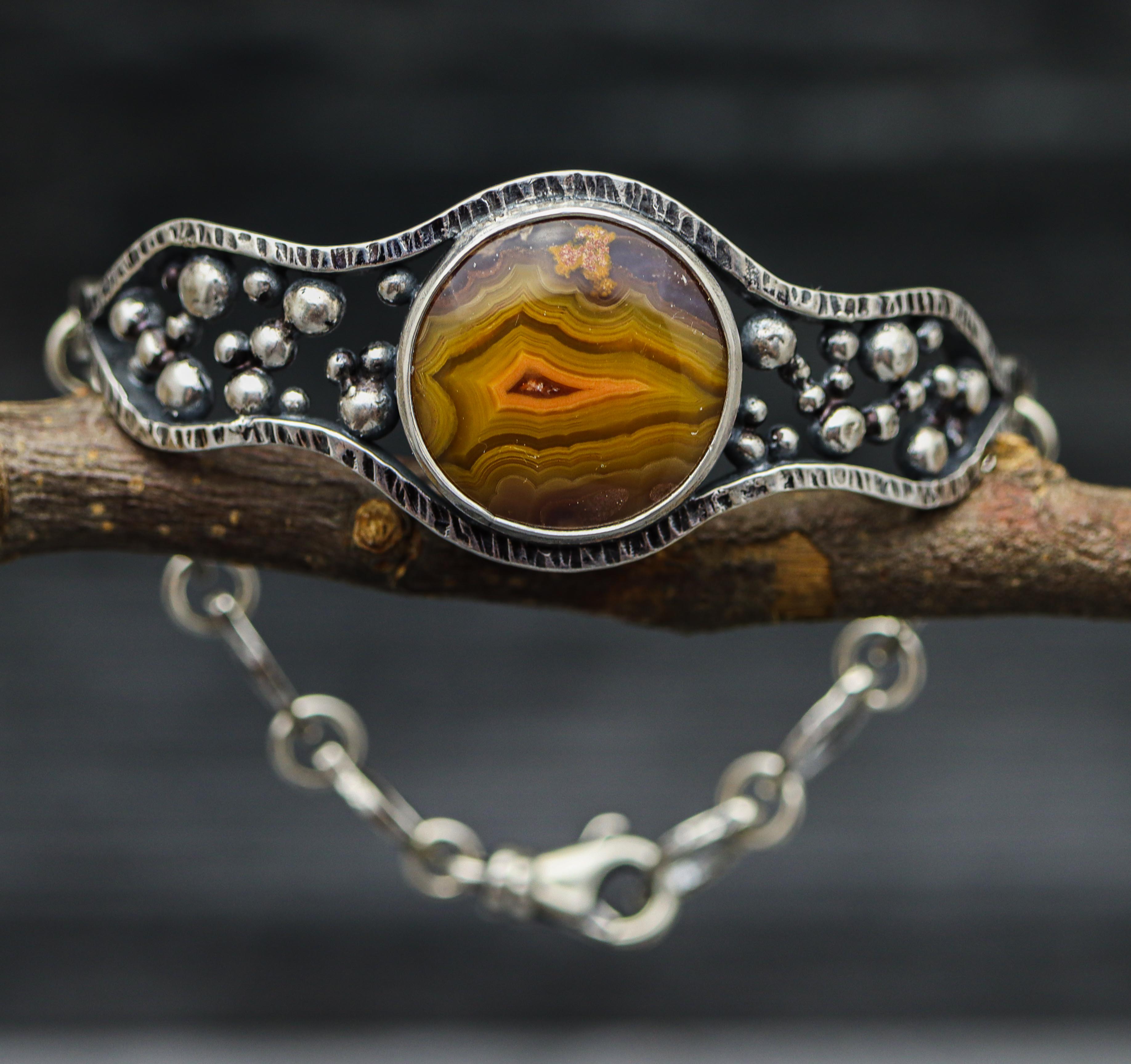 A sterling silver cuff bracelet with a hybrid design, featuring a central laguna agate stone surrounded by granulated details, displayed on a branch against a dark background.