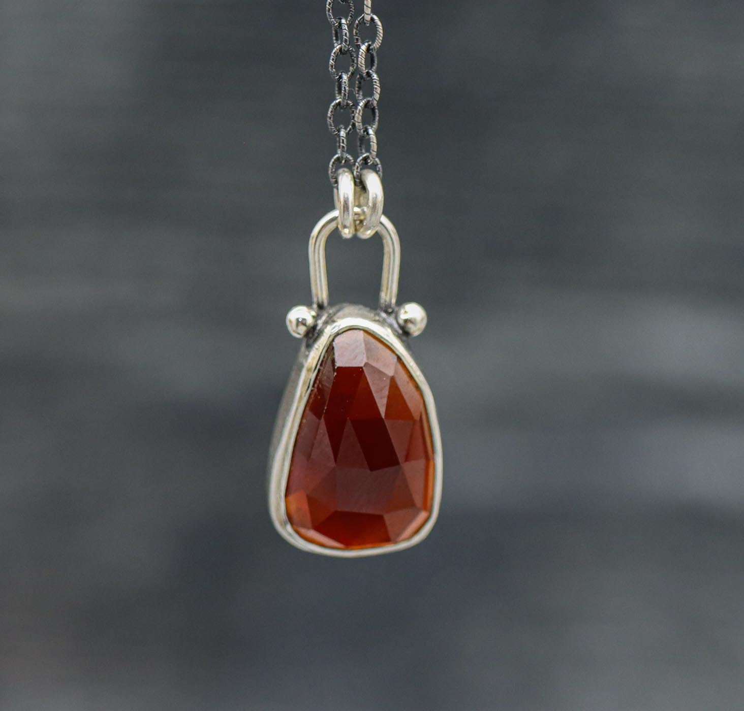 A hessonite garnet pendant necklace in sterling silver with small silver ball accents, displayed against an oxidized background.