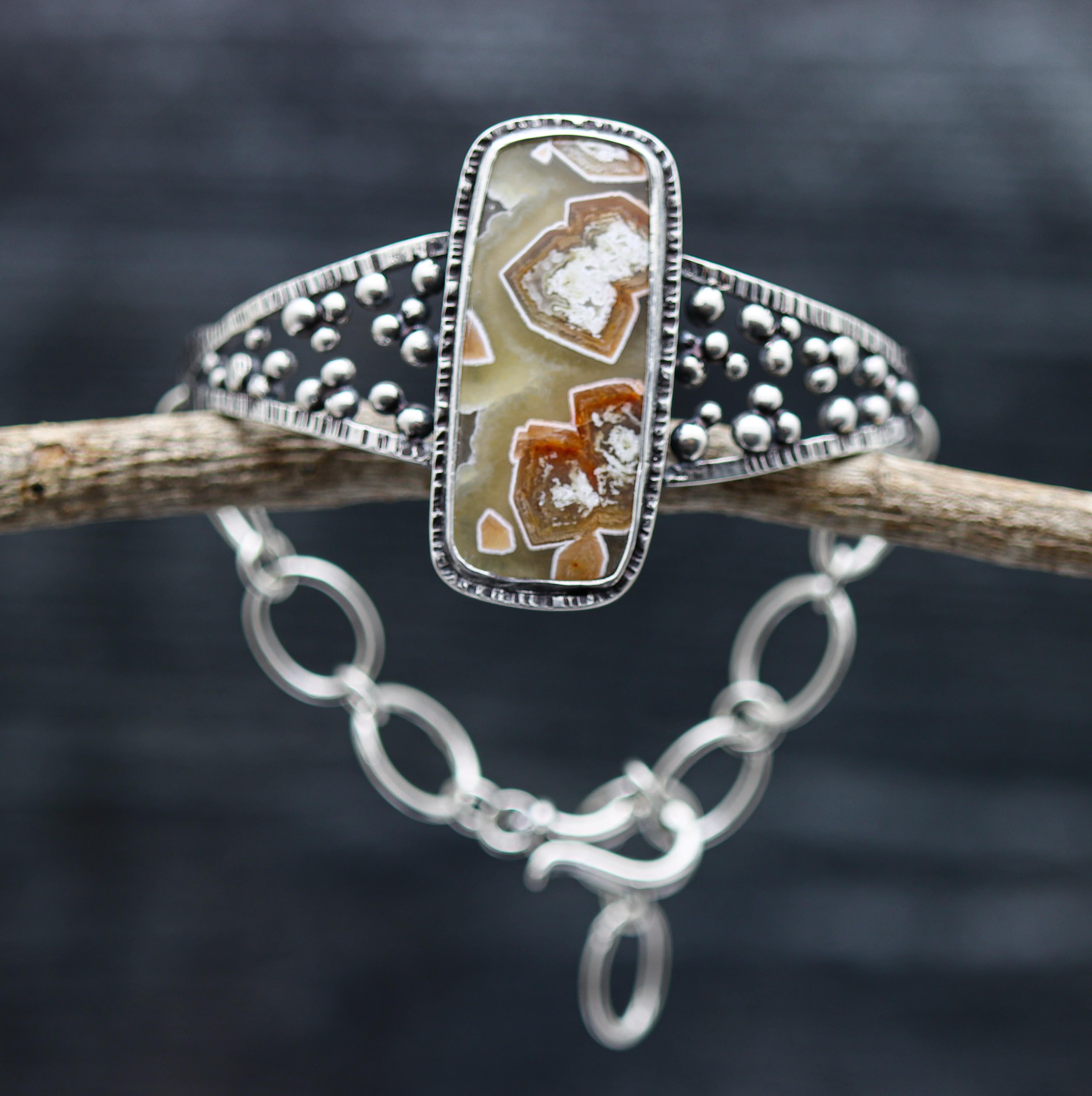 A sterling silver cuff bracelet with a hybrid design, featuring a central laguna agate stone surrounded by silver granules, displayed on a dark background.