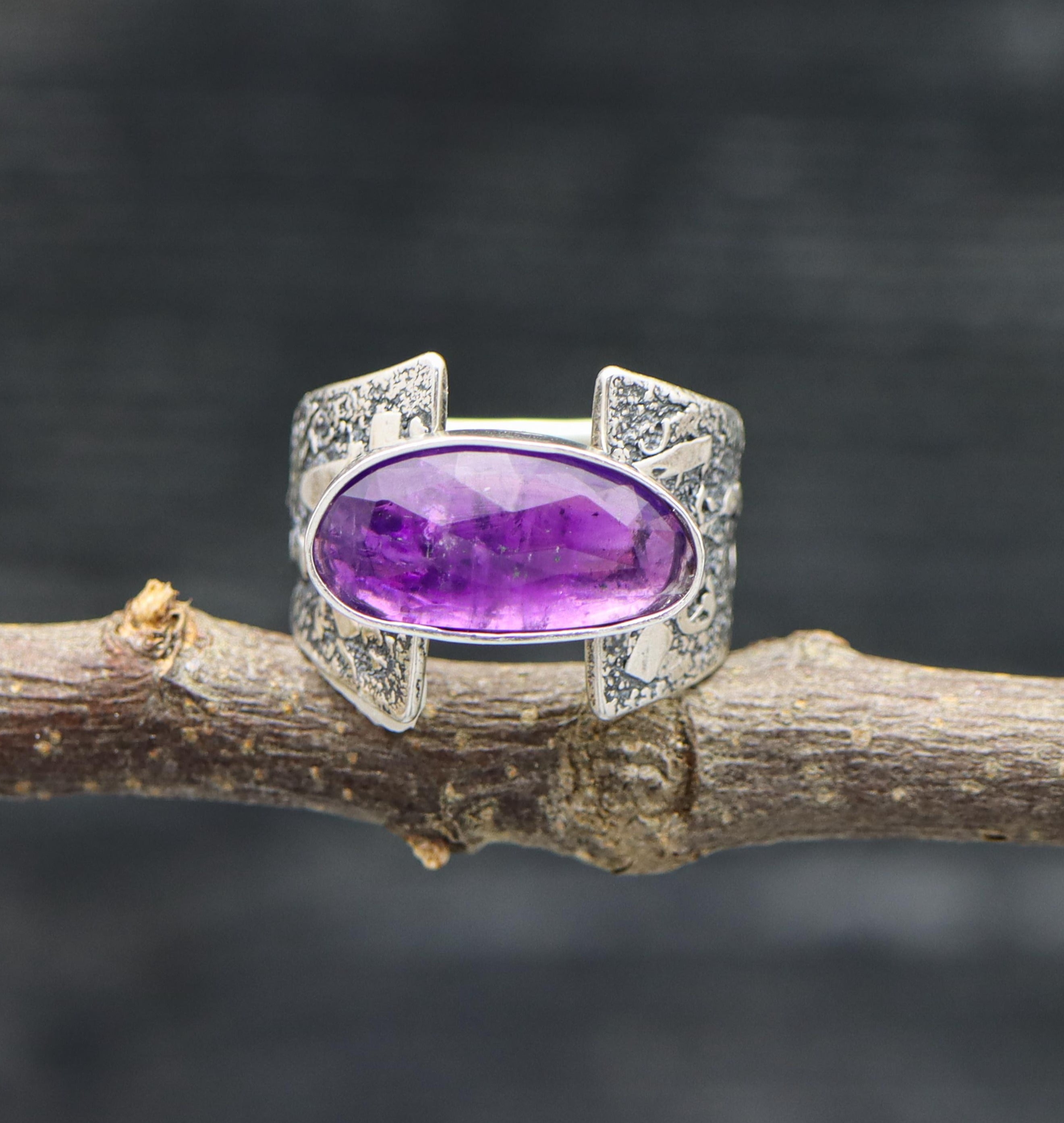 A sterling silver ring with a large purple amethyst stone set in a silver setting, displayed on a rustic branch background.