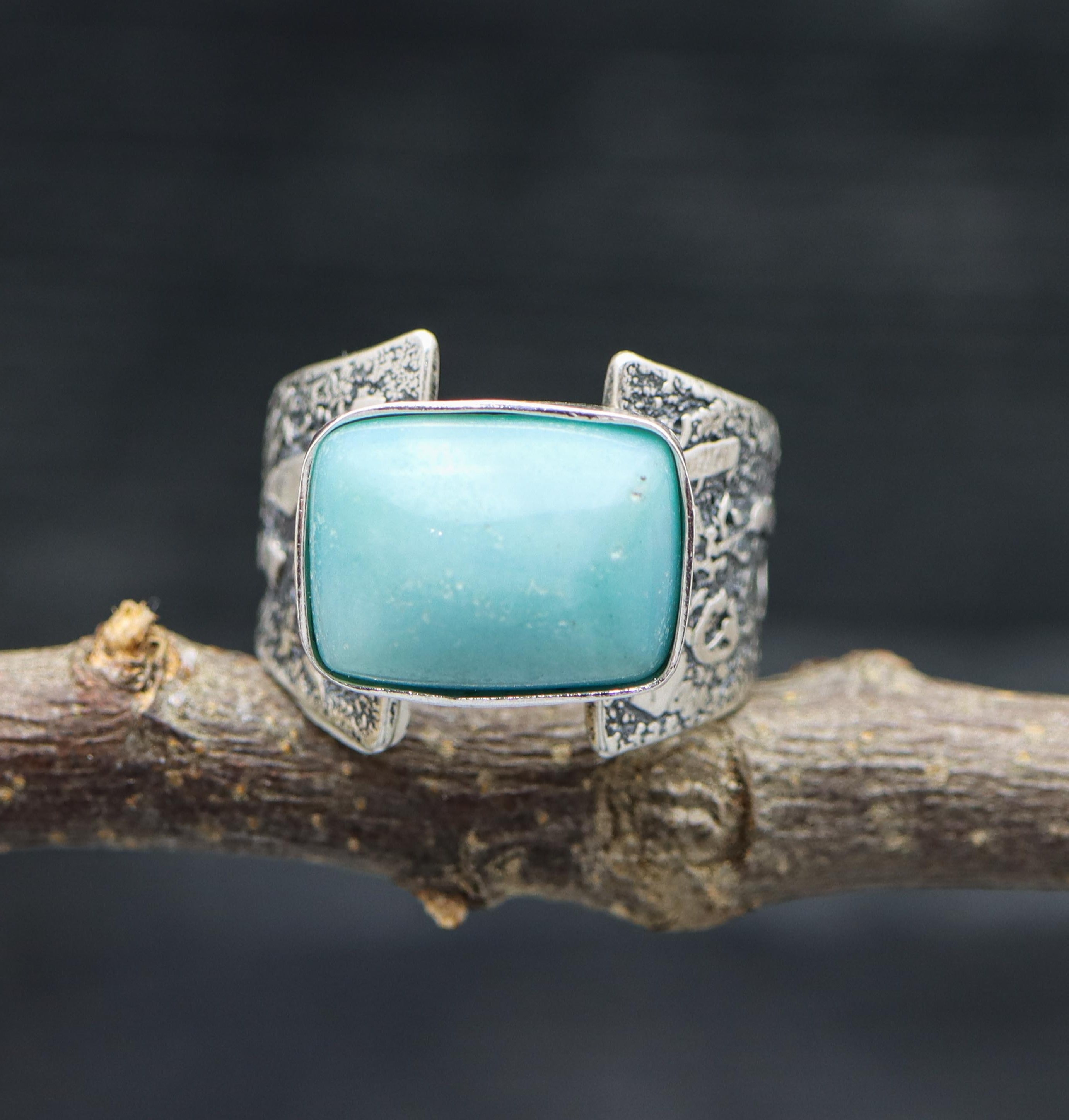 A sterling silver ring with a blue amazonite stone set on a wide band, showcased on a rustic branch background.