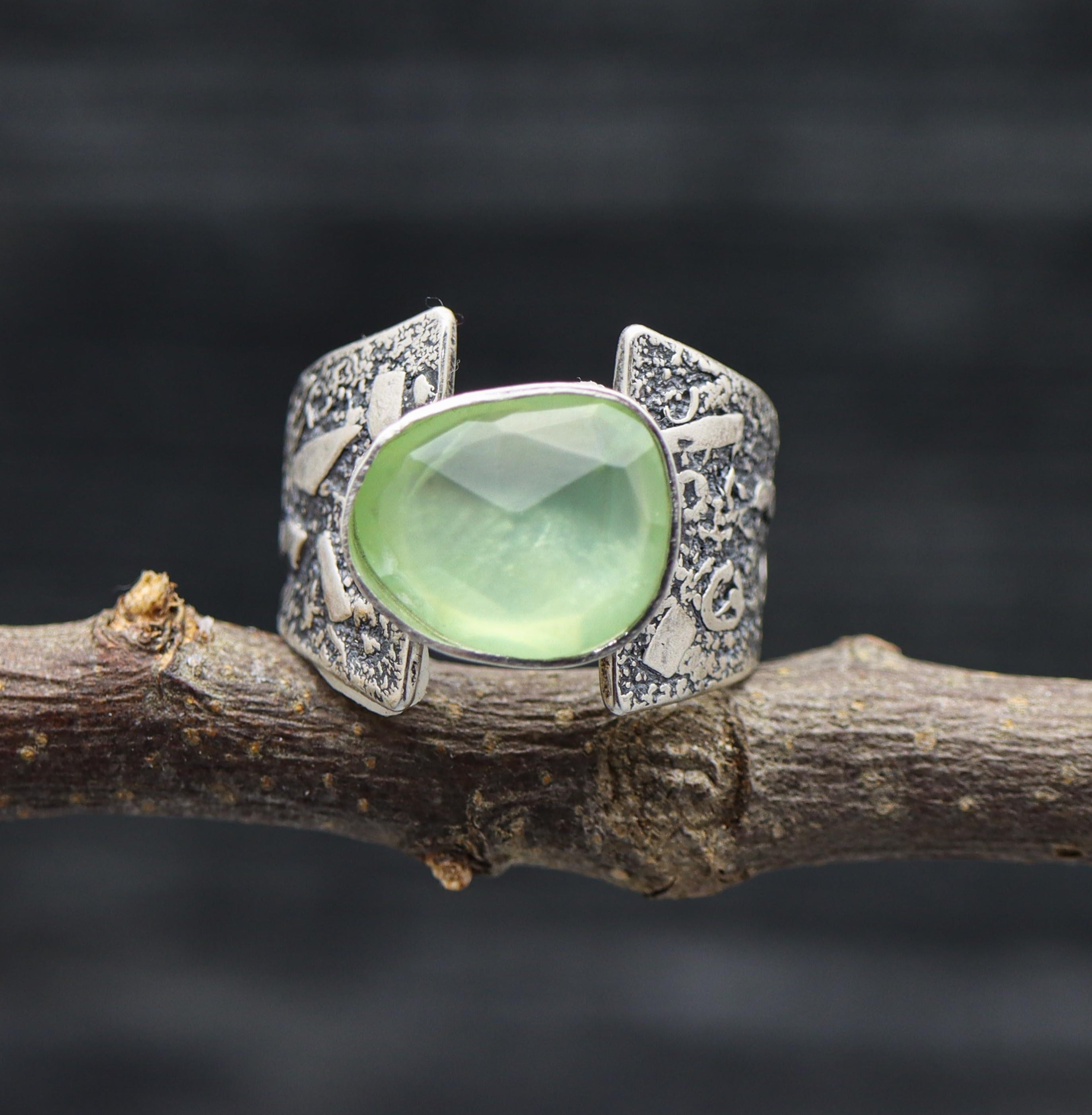 A sterling silver ring with a faceted rose cut green prehnite stone, displayed on a dark branch against a dark background.