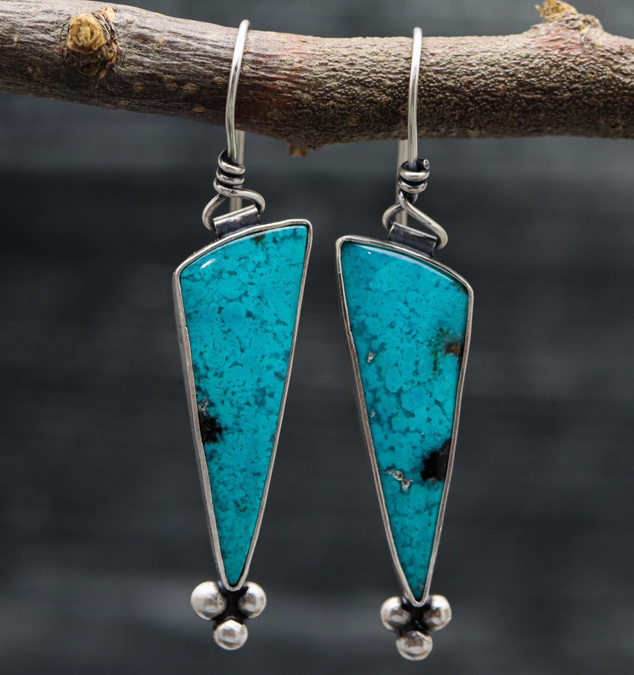 A pair of sterling silver dangle earrings with blue chrysocolla stones and black tenorite accents, displayed on a branch.