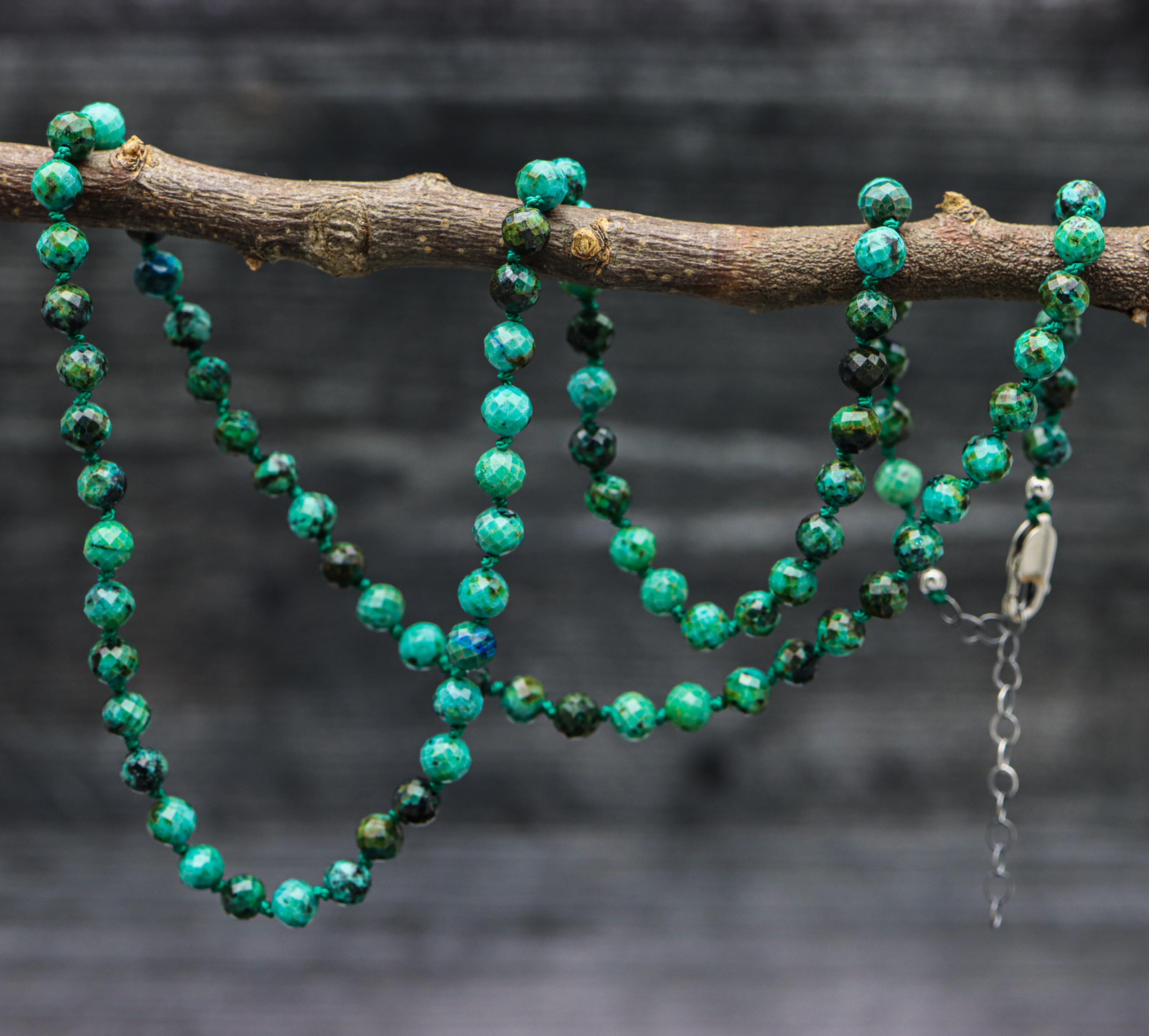 A hand knotted strand of chrysocolla beads on dark green thread with sterling silver metal, displayed on a branch.