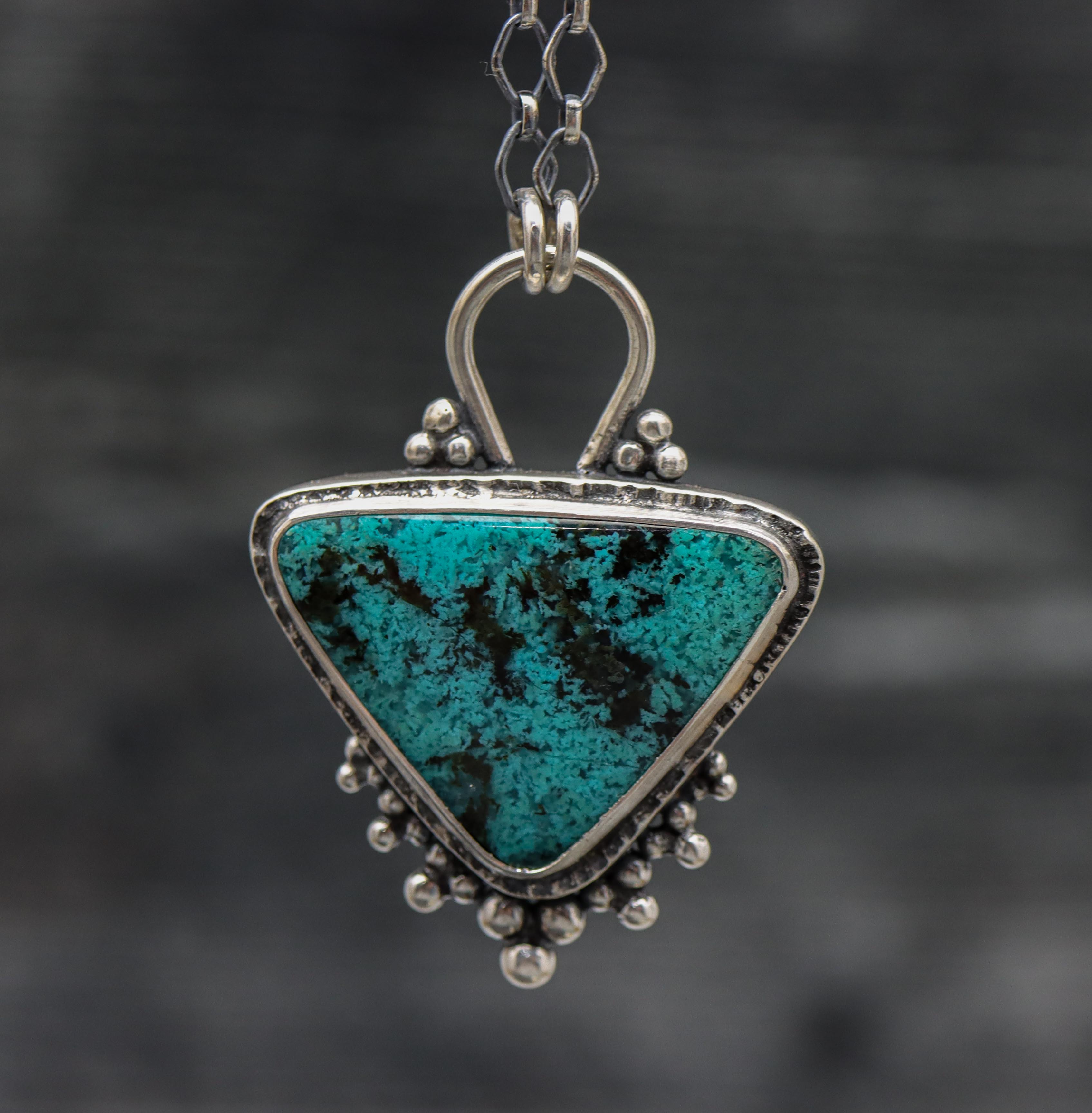 A sterling silver pendant featuring a turquoise blue chrysocolla gemstone with black speckles, set in a sterling silver bezel, attached to a diamond link chain with a lobster claw clasp.
