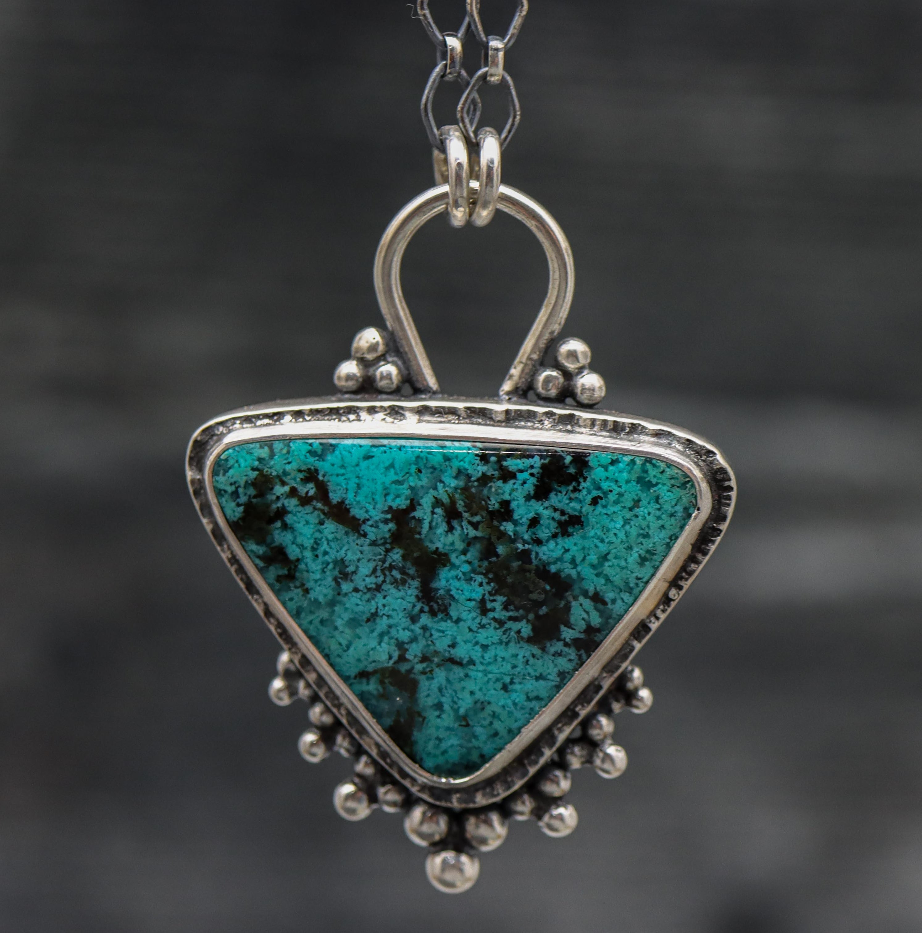 A sterling silver pendant featuring a turquoise blue chrysocolla gemstone with black speckles, set in a sterling silver bezel, attached to a diamond link chain with a lobster claw clasp.