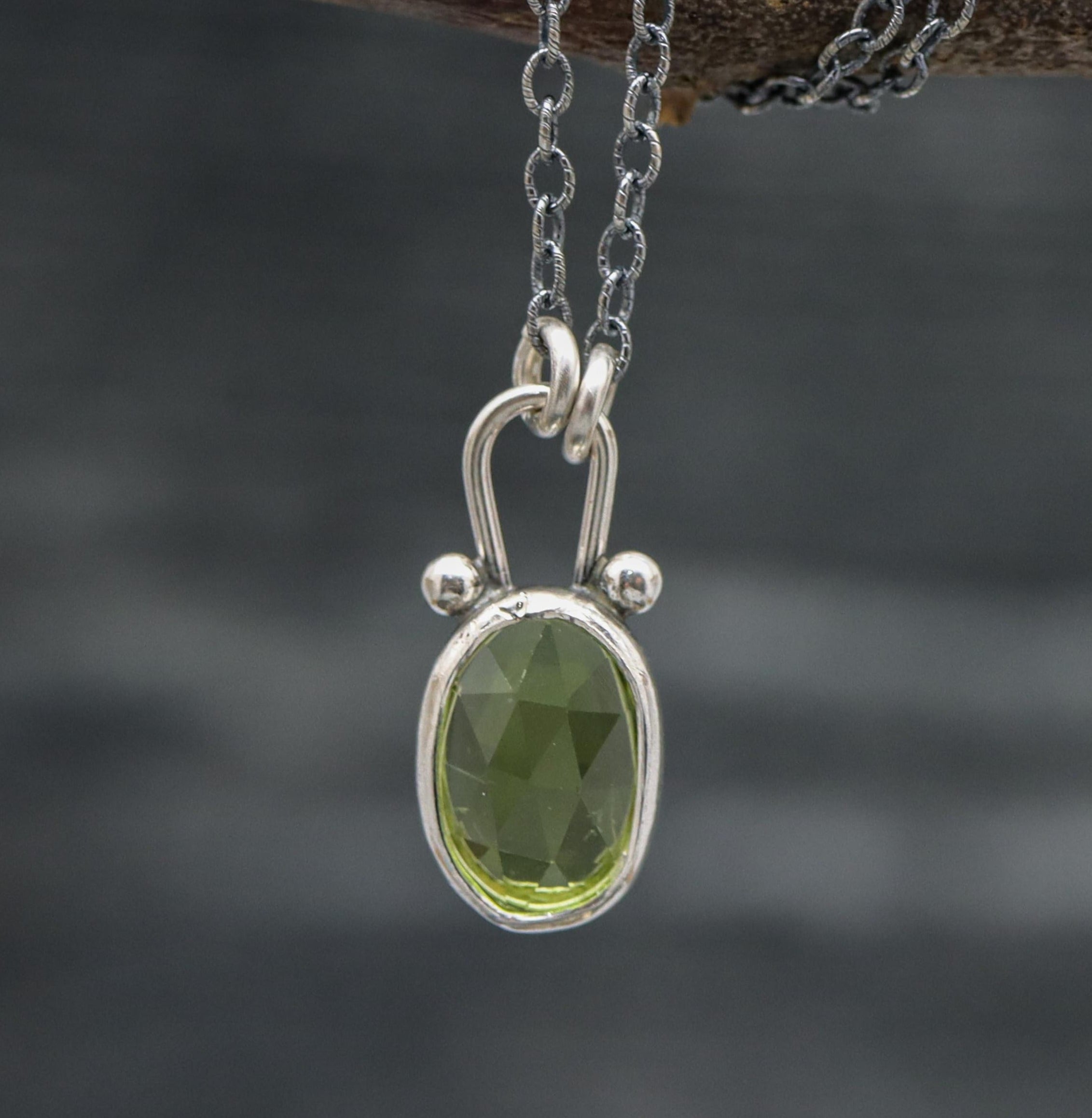A sterling silver pendant necklace with a faceted rose cut green peridot, displayed on a branch.