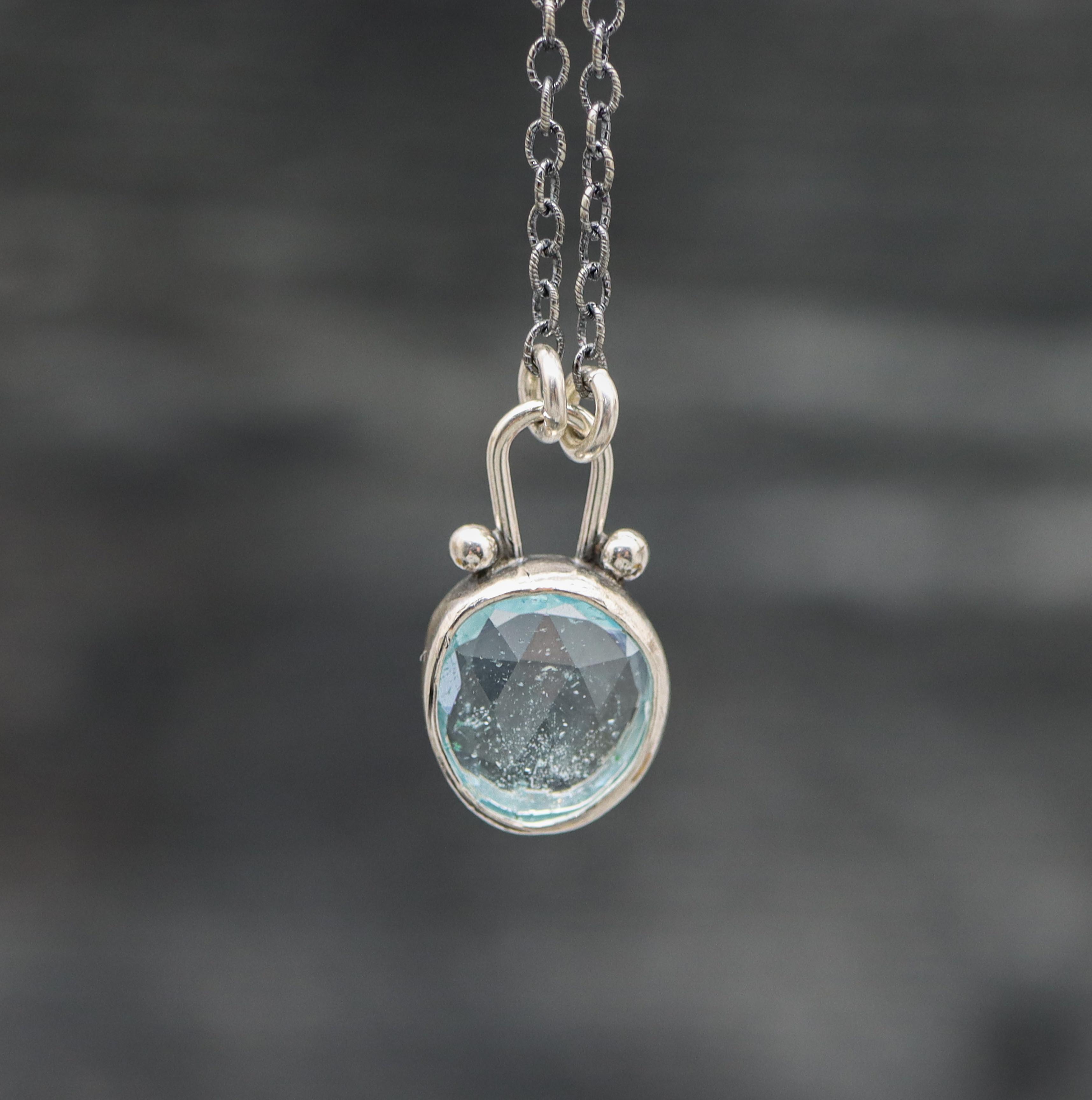 A sterling silver pendant necklace with a faceted rose cut blue topaz centerpiece, surrounded by small silver ball accents, on an oxidized cable link chain.