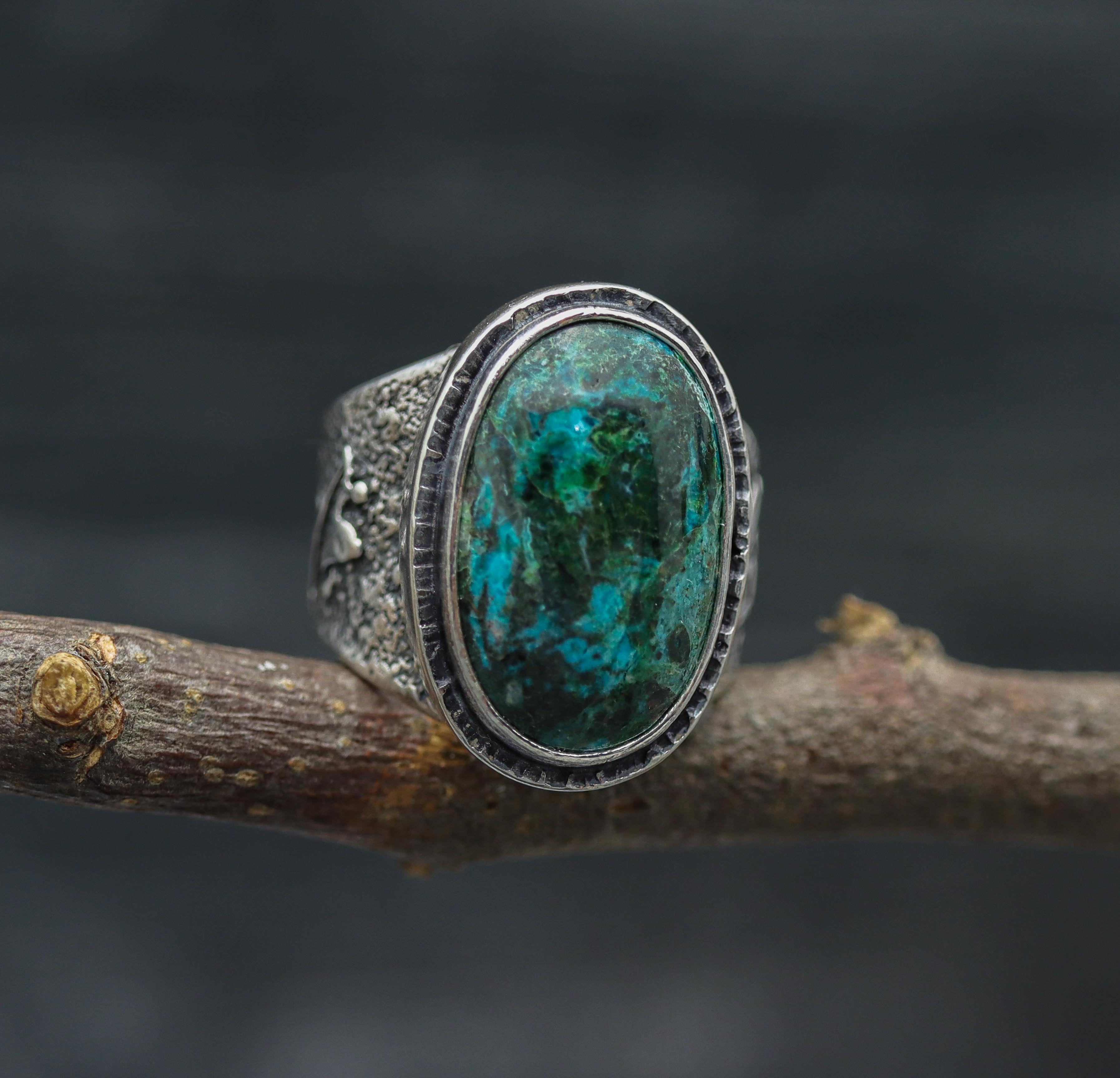 A sterling silver wide band ring with a chrysocolla in quartz stone set in a double bezel setting, displayed on a rustic wooden branch.