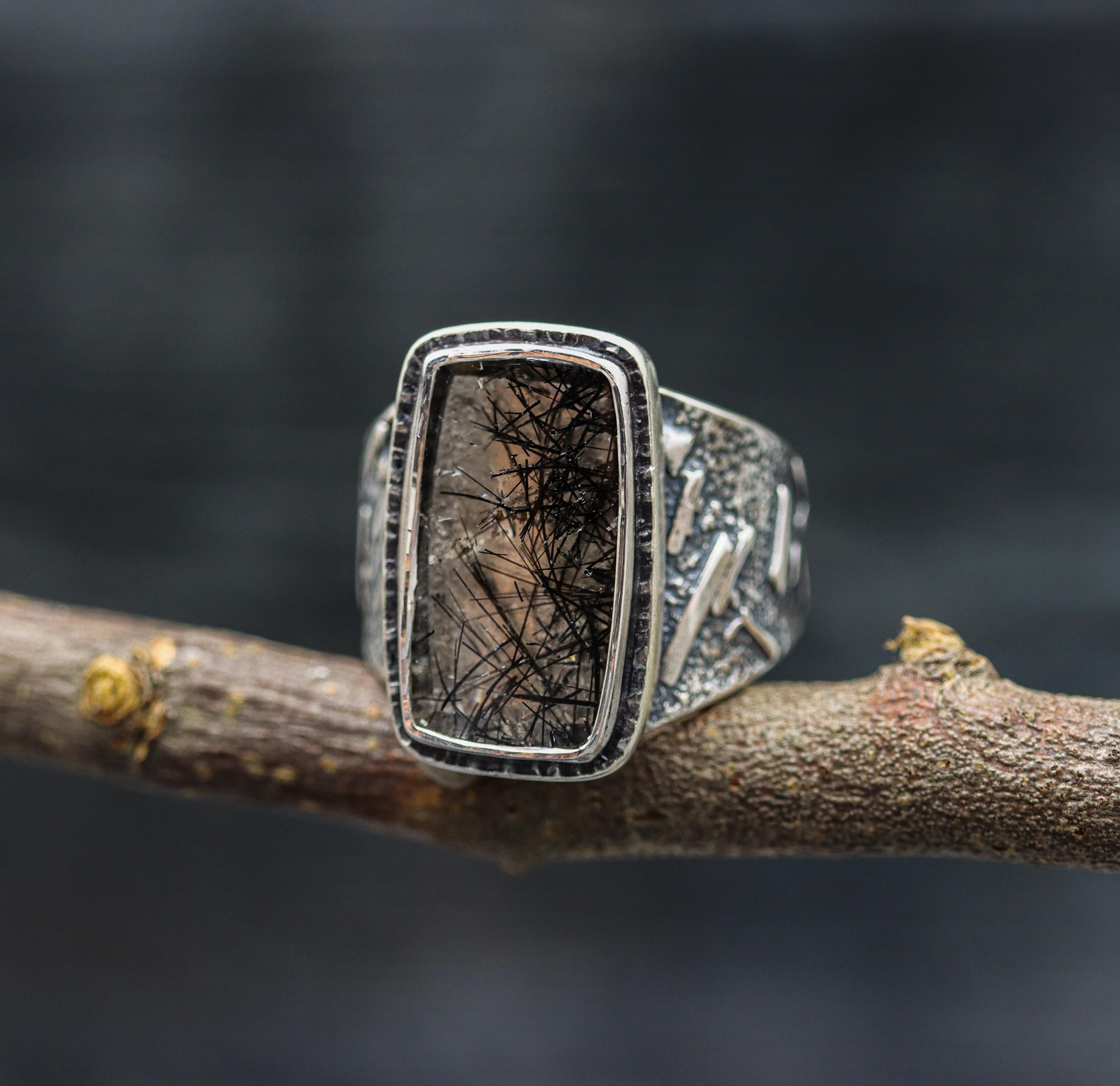 A wide band sterling silver ring with a black tourmalinated quartz centerpiece, set in a double bezel setting, displayed on a rustic wooden branch.