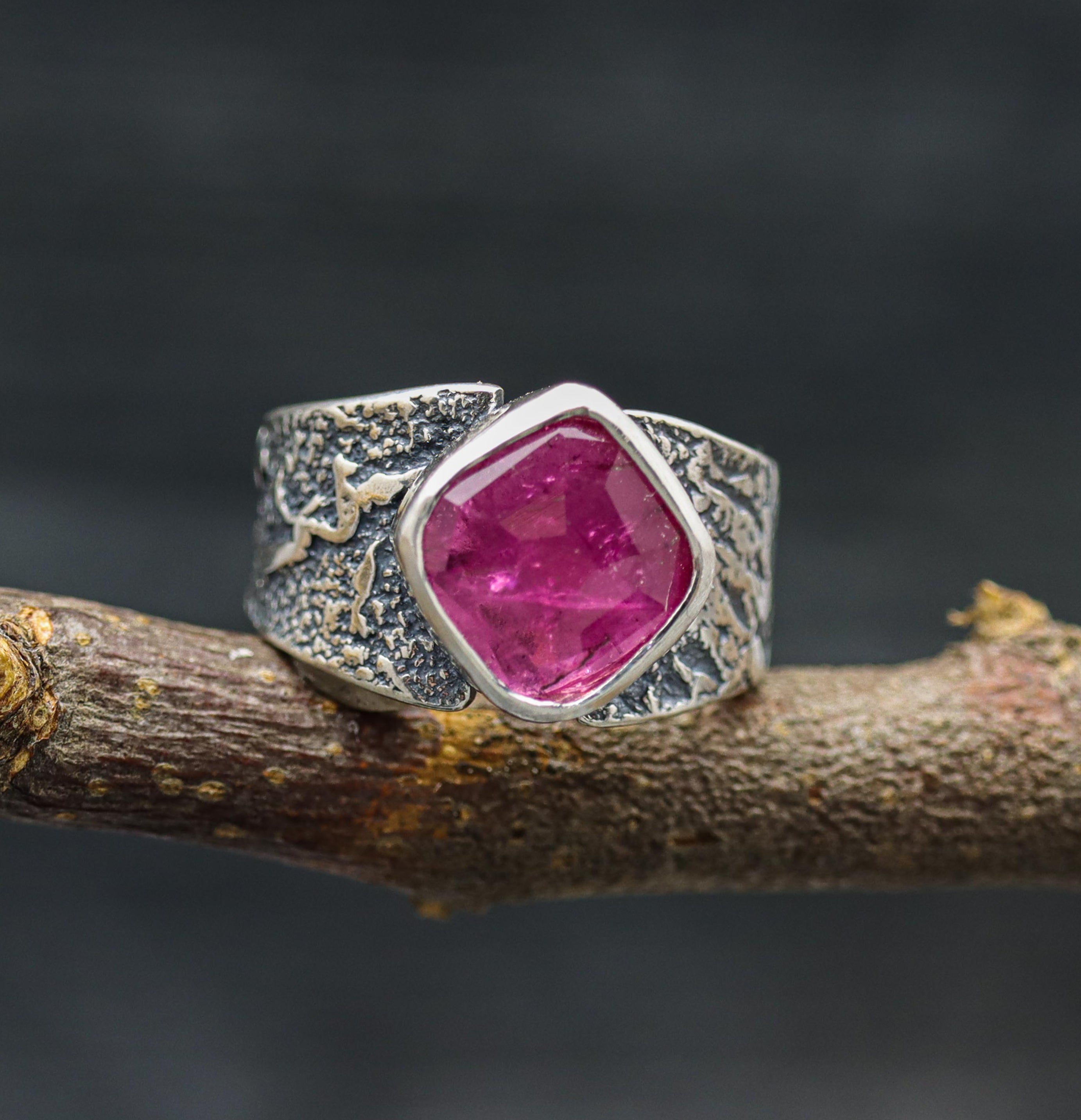 A wide band sterling silver ring with a bold pink tourmaline centerpiece, displayed on a branch against a dark background.