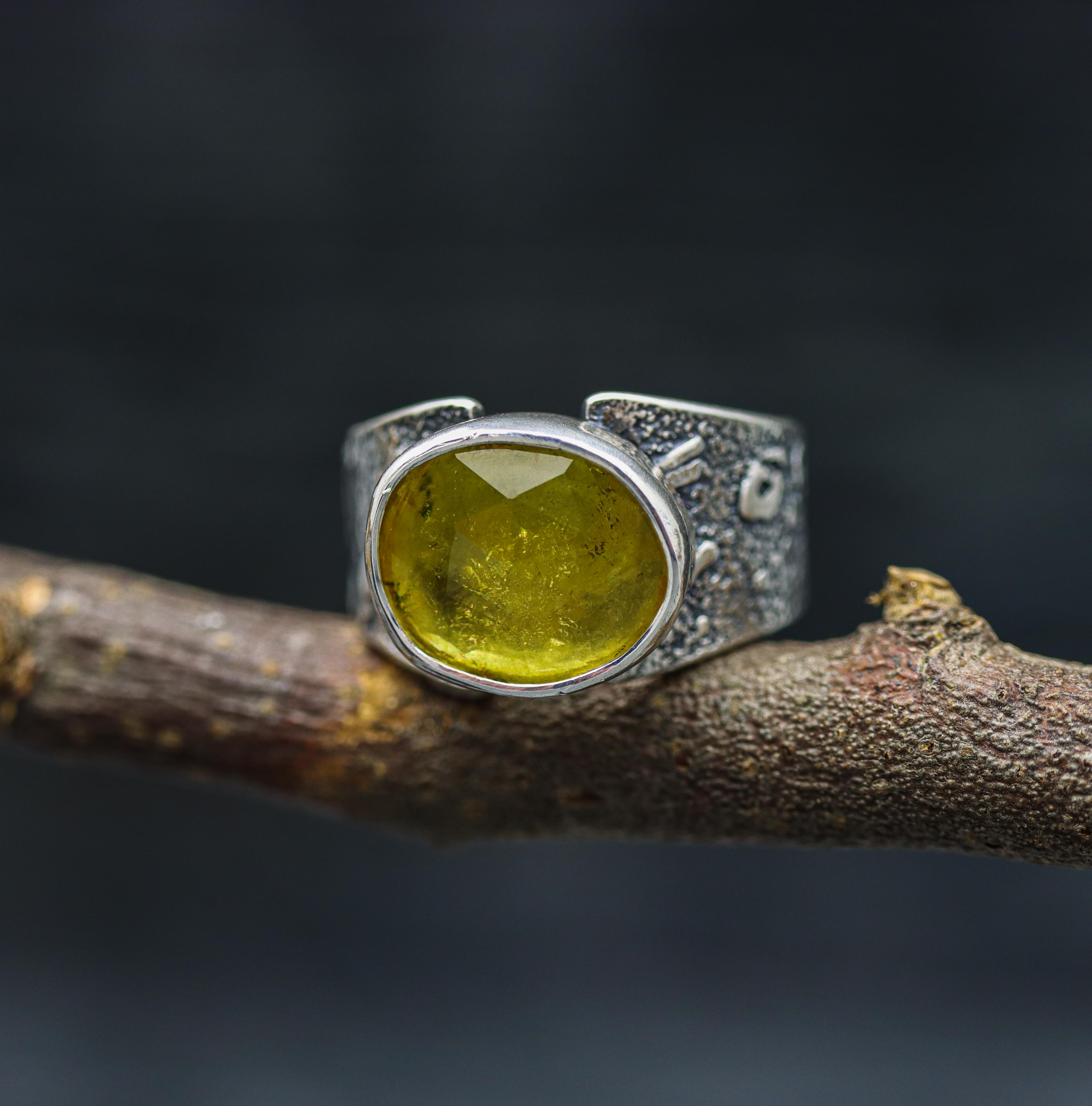 A yellow tourmaline gemstone ring with a wide band, set in a sterling silver setting, displayed on a rustic wooden branch.