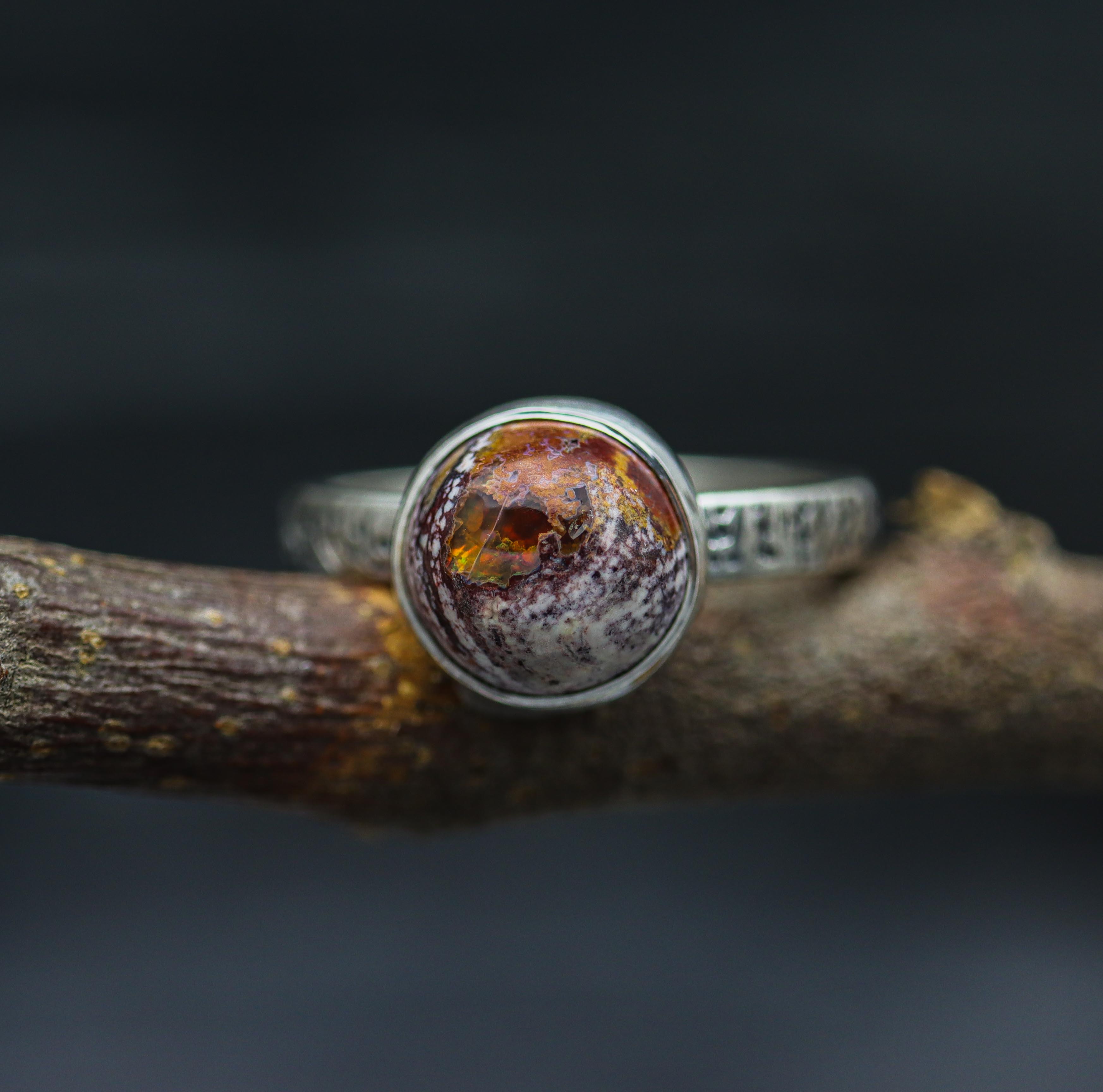 A sterling silver ring with a Mexican fire opal gemstone set on a textured band, displayed on a branch.