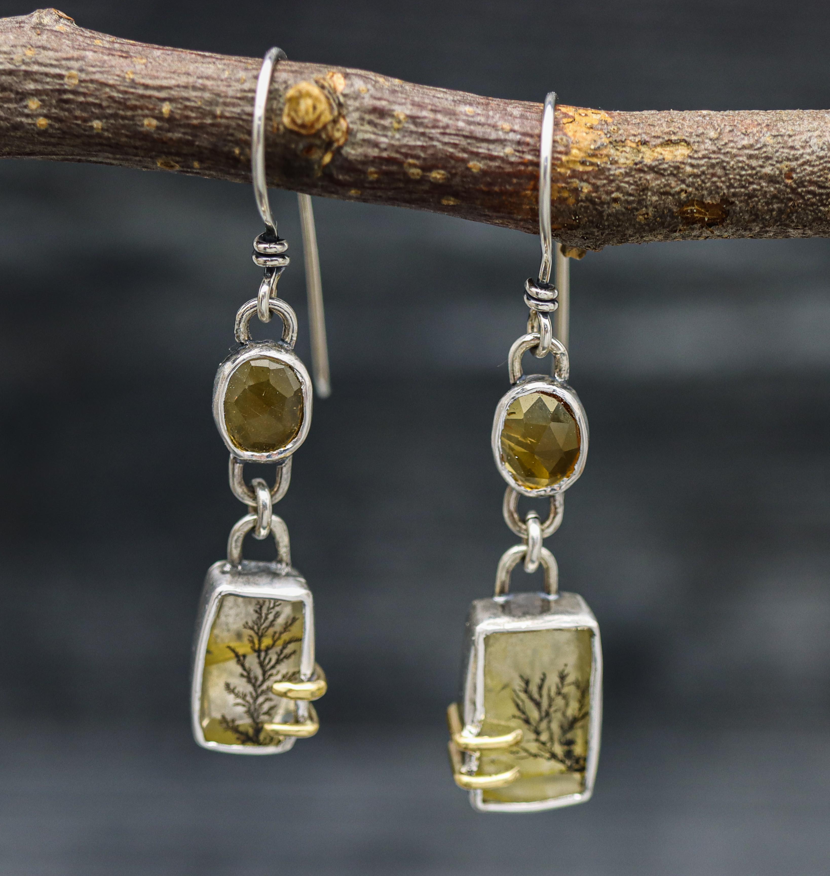 A pair of sterling silver dangle earrings with both featuring a dendritic quartz and citrine, both with gold prongs, displayed on a tree branch.