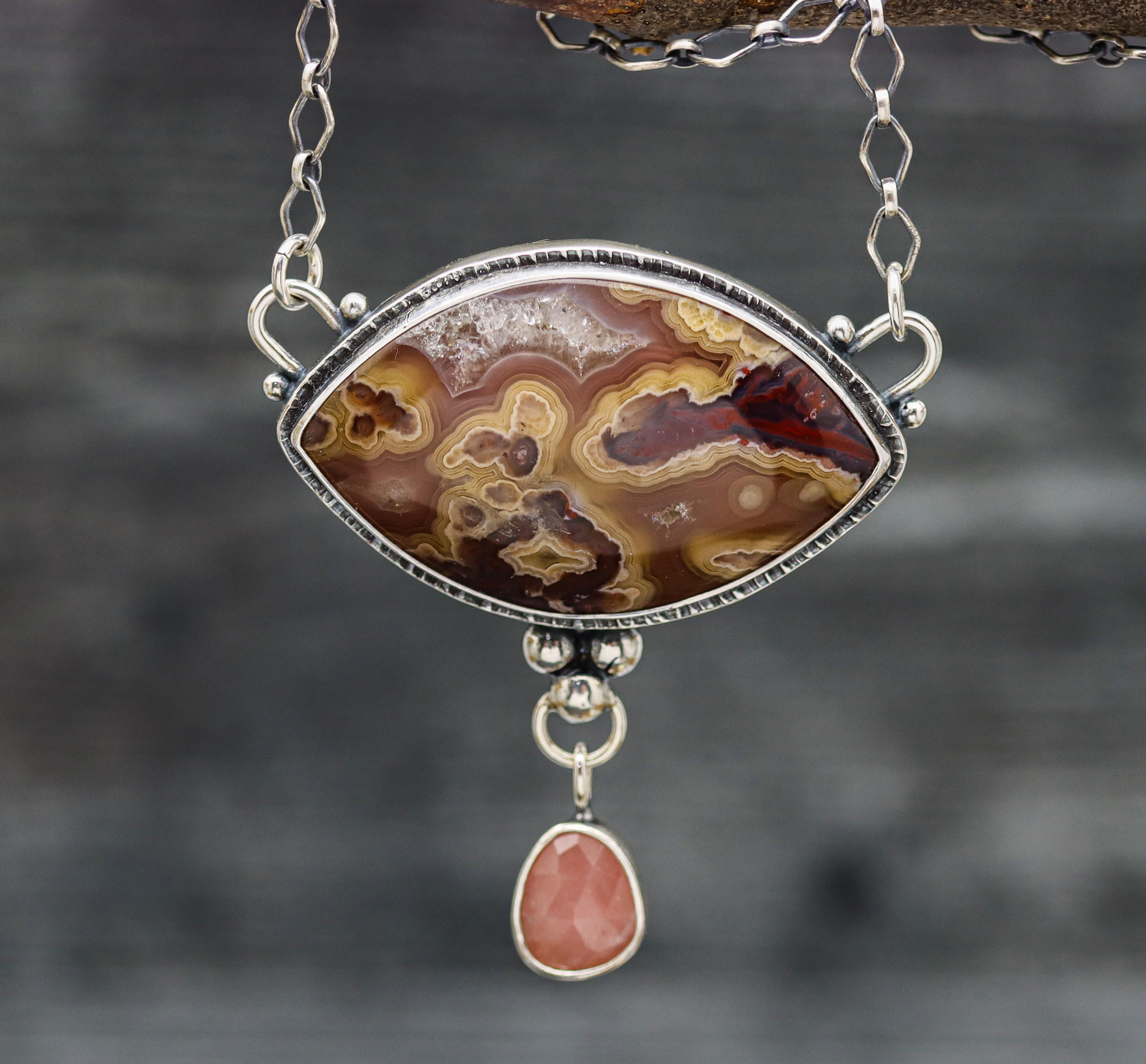 A sterling silver necklace featuring a laguna agate gemstone with a druzy center and a guava quartz accent, displayed on a silver chain against a textured background.