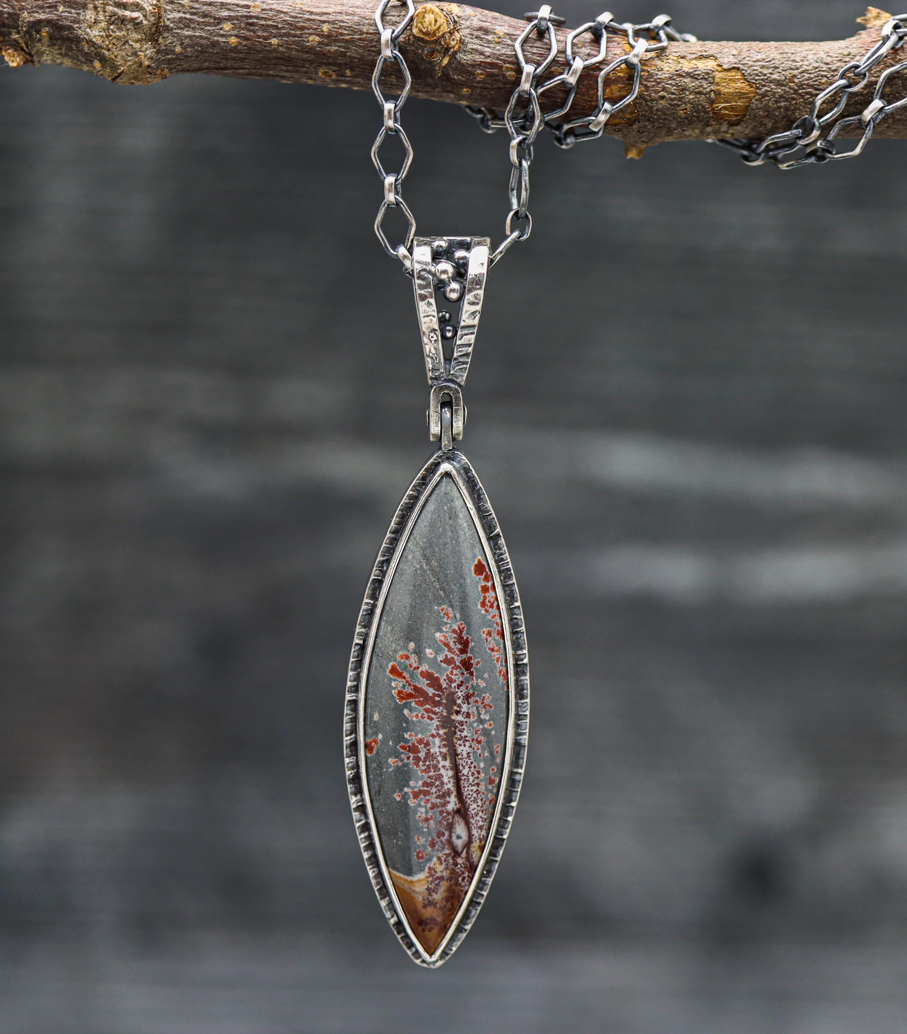 A sterling silver necklace featuring a polished Sonoran dendritic rhyolite jasper gemstone with a blue-grey color and red dendrites, displayed against a natural outdoor background.