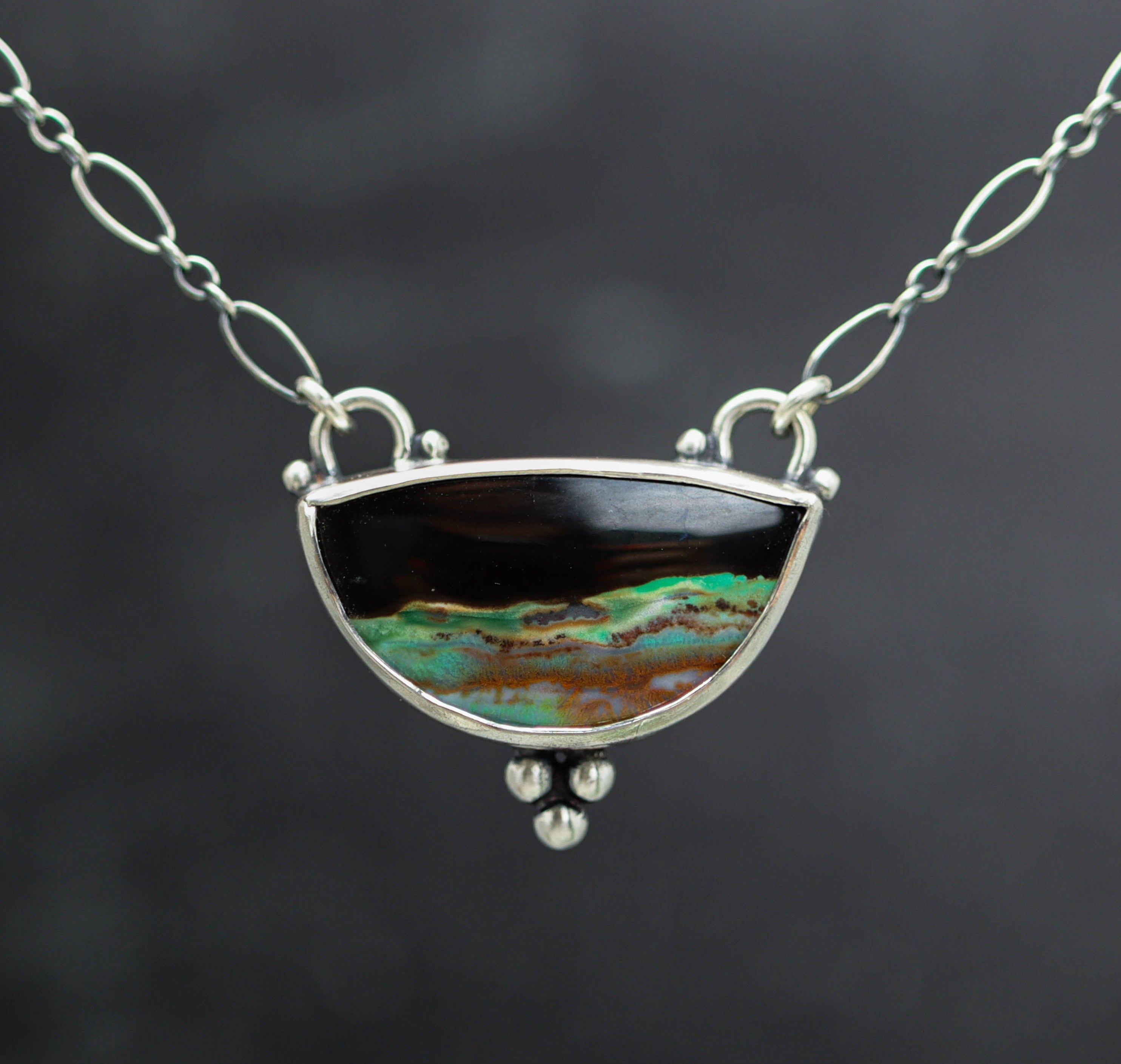 A silver link chain necklace featuring a semi-circular pendant with a black top section and a bottom section displaying a vibrant green, turquoise, and brown marbled pattern, set against a dark gray background.