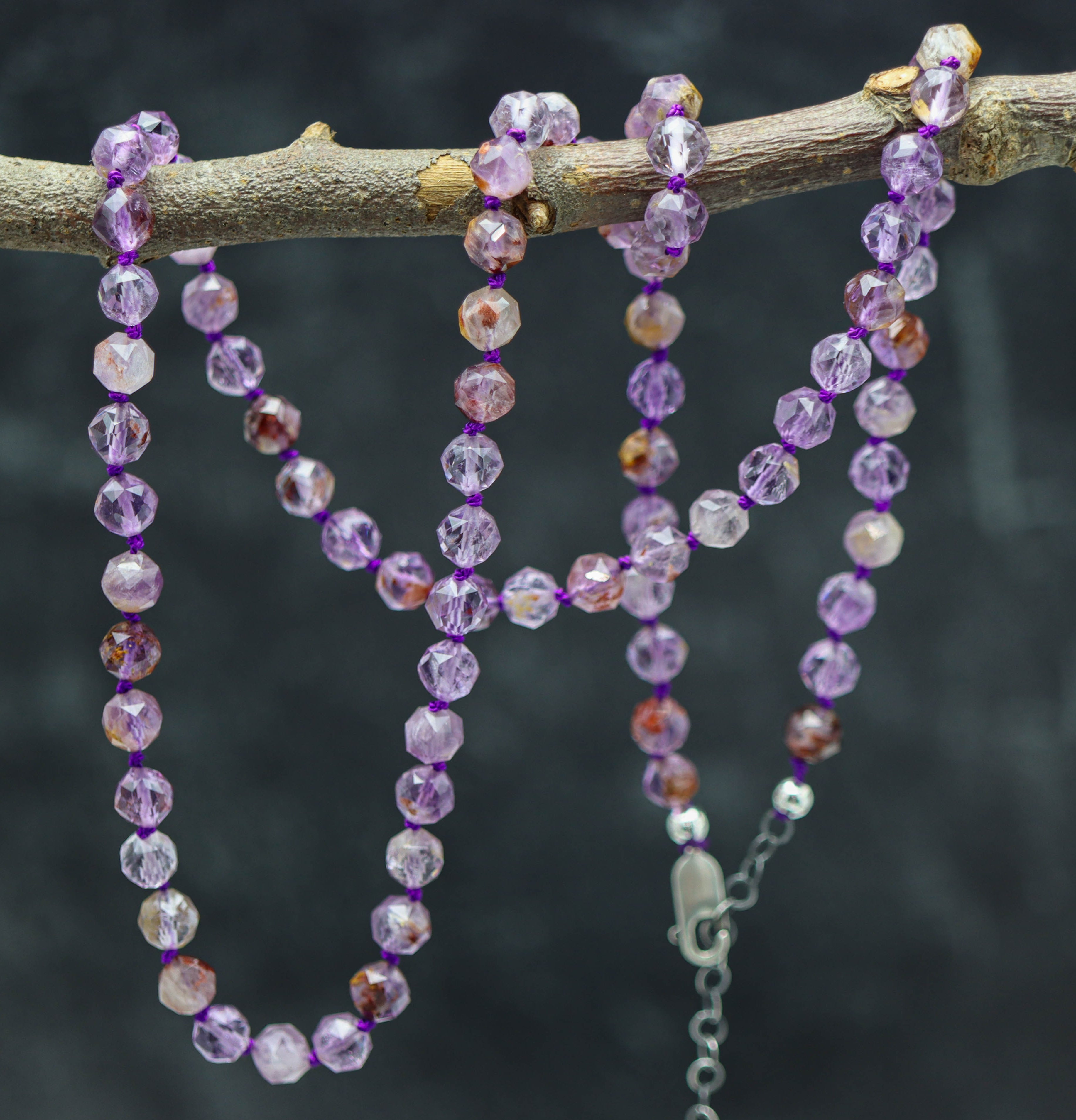 A hand knotted necklace made of purple super seven beads and sterling silver, displayed on a branch.