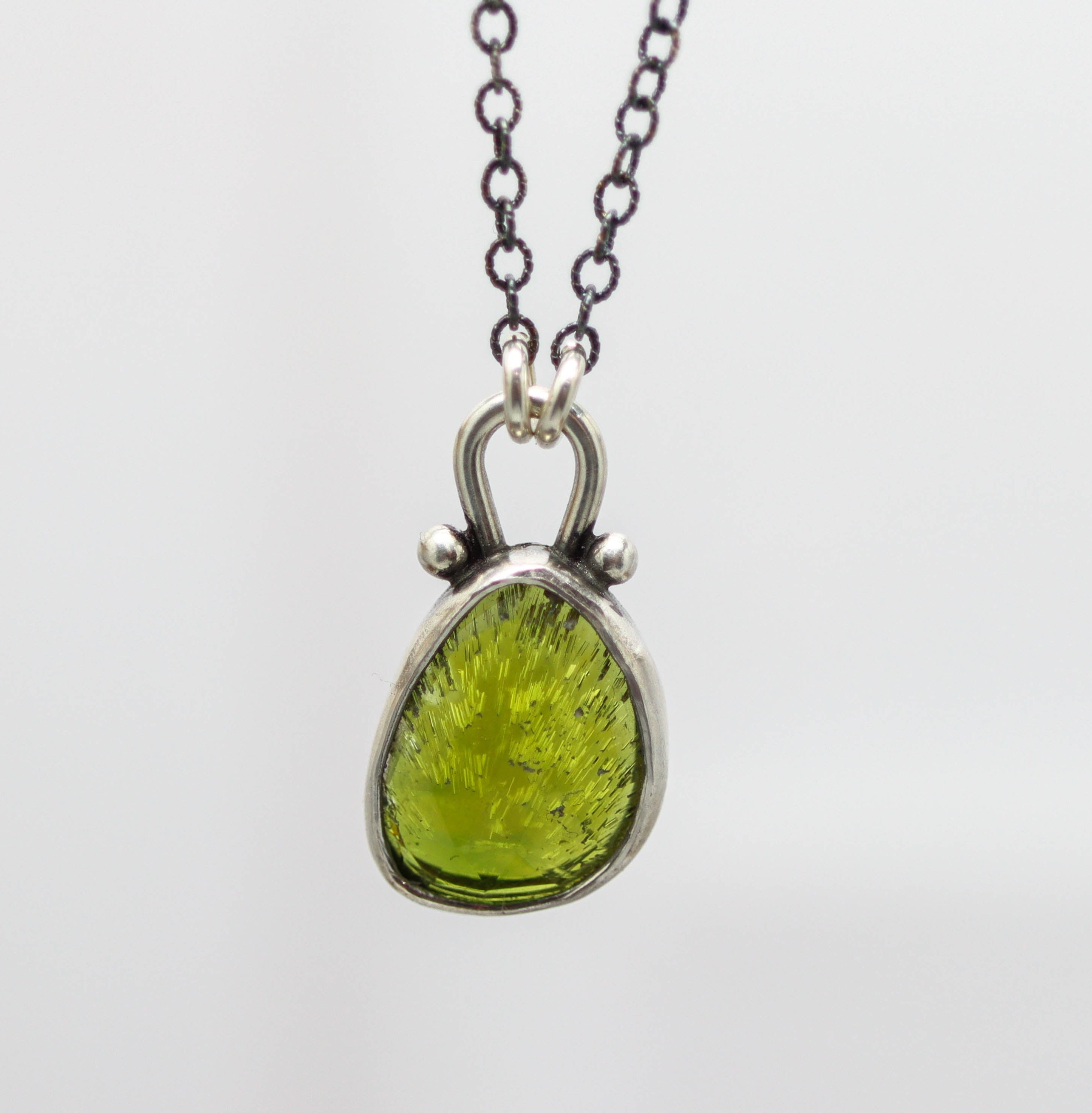A sterling silver pendant necklace featuring a rose-cut green tourmaline centerpiece with metallic detailing and a lobster claw closure.