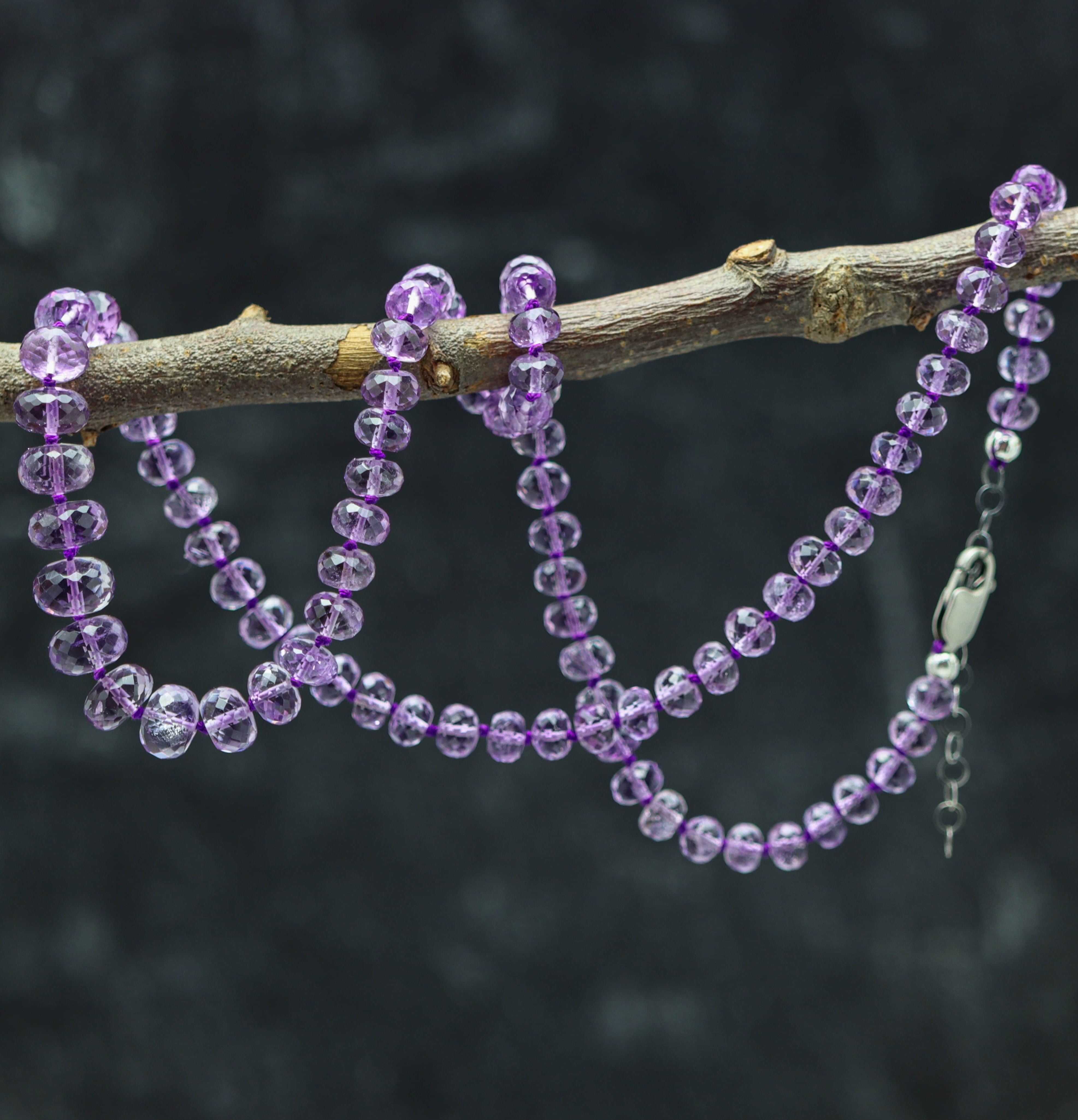A sterling silver necklace with a series of graduated amethyst hand knotted beads, displayed on a branch against a dark background.
