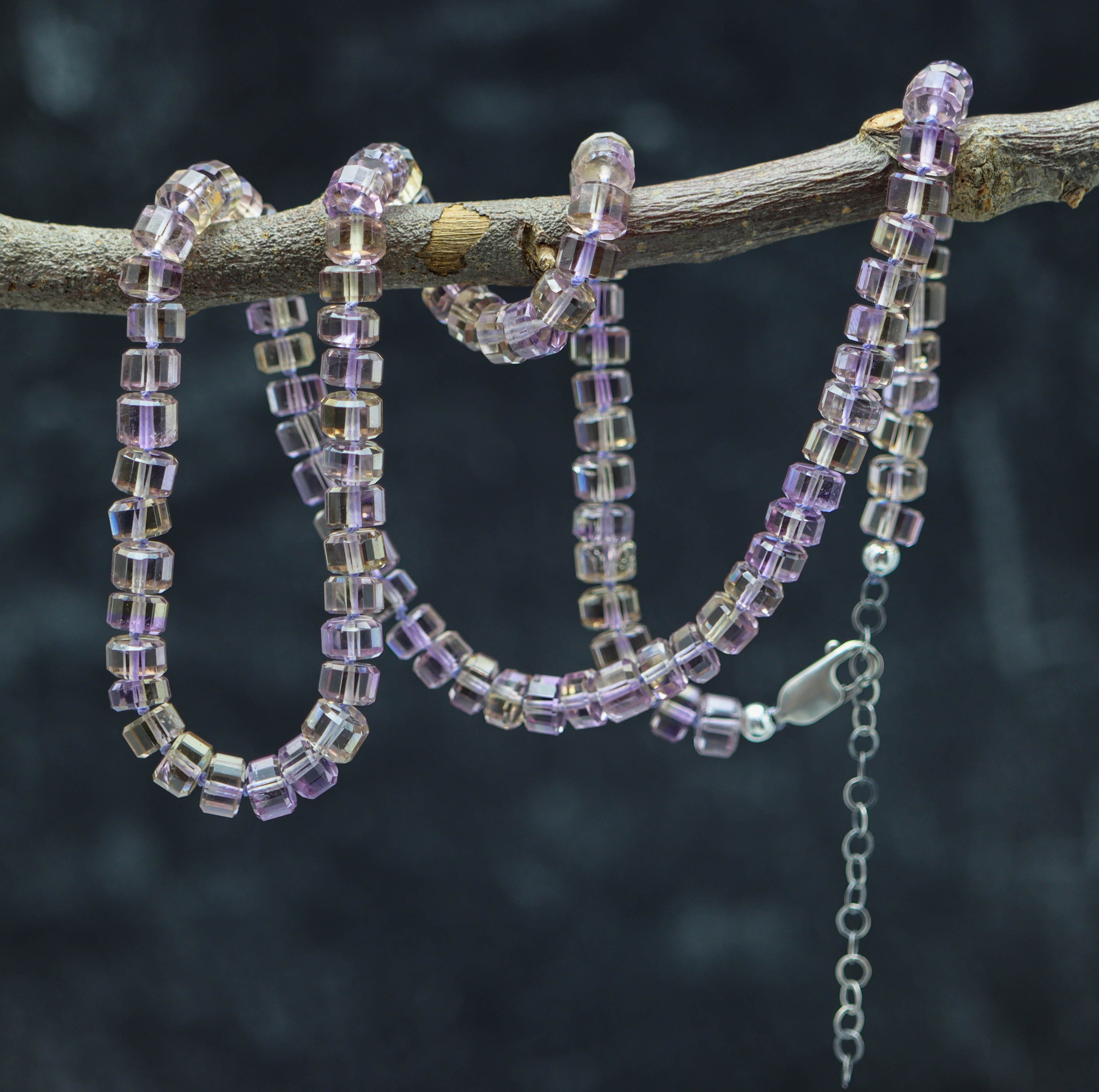 A hand knotted bead necklace with sterling silver faceted barrels, featuring a blend of purple and clear beads, displayed on a branch against a dark background.