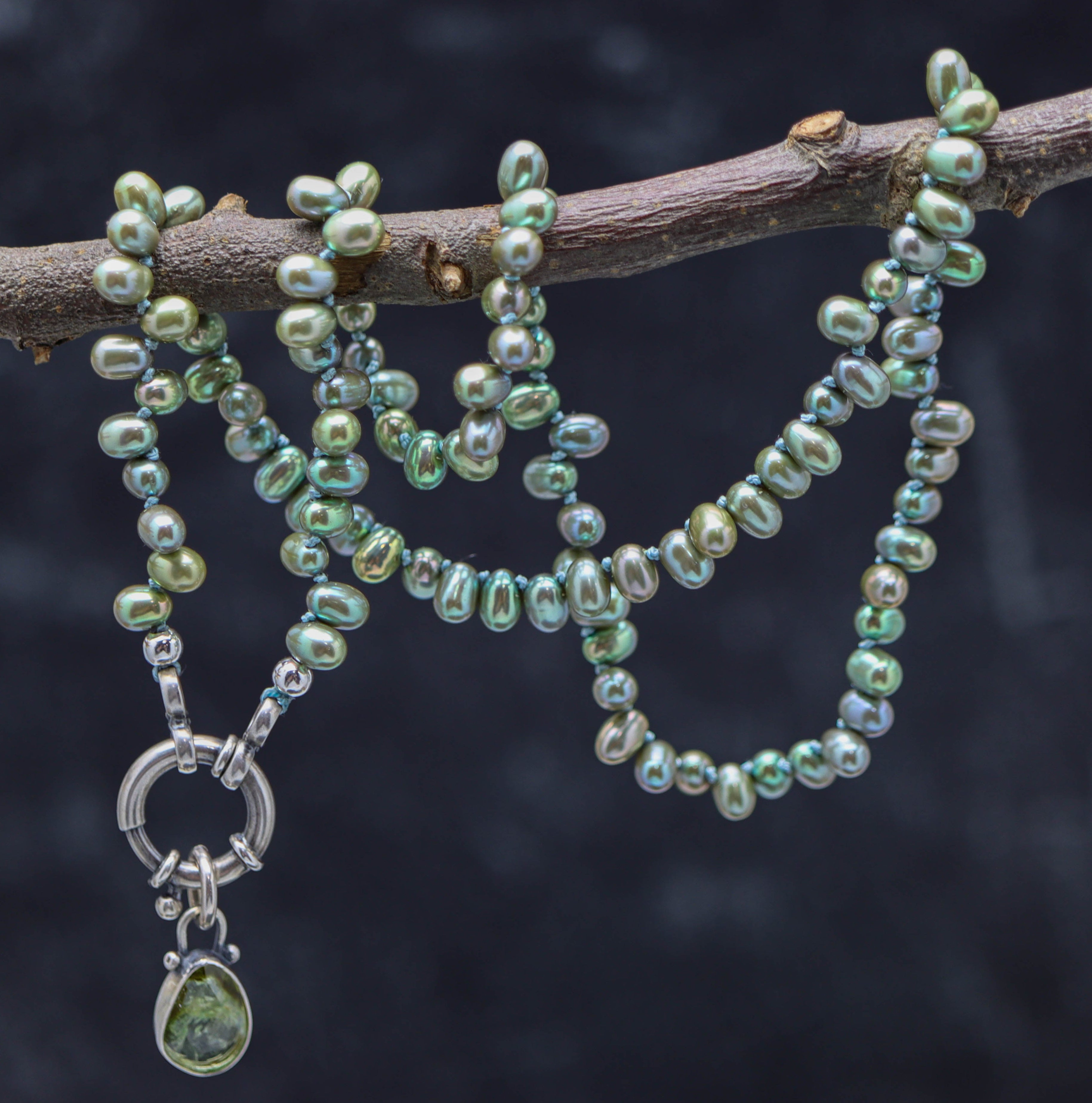 A sterling silver necklace featuring hand knotted beads with green freshwater pearls and a tourmaline pendant, displayed on a branch against a blurred background.