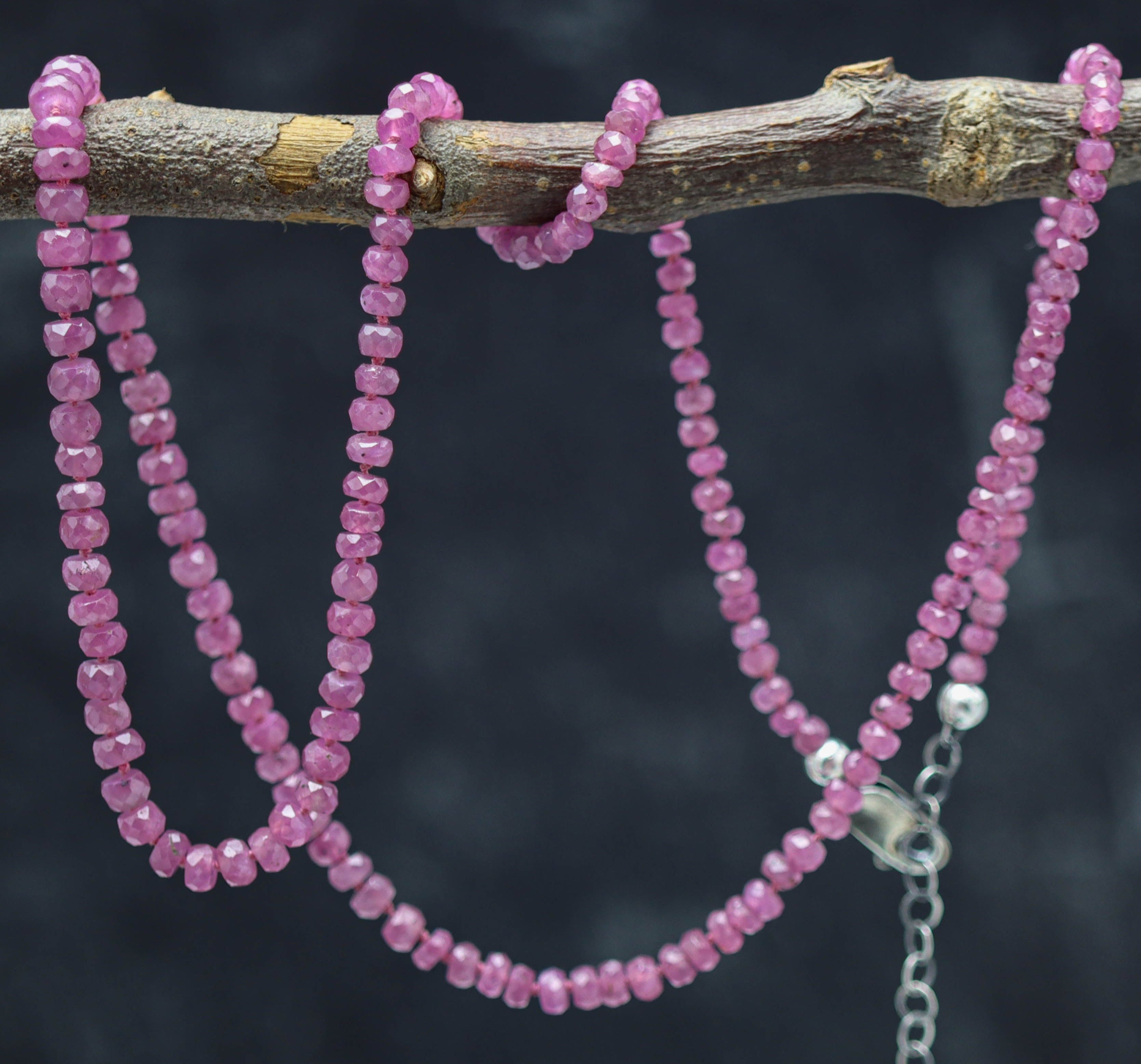 A hand knotted necklace made of pink ruby beads, strung on silk thread with a silver clasp, displayed on a branch.