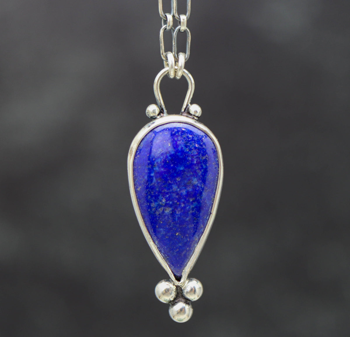 A sterling silver pendant necklace with a blue lapis lazuli gemstone centerpiece and small silver ball accents, featuring a lobster claw closure.