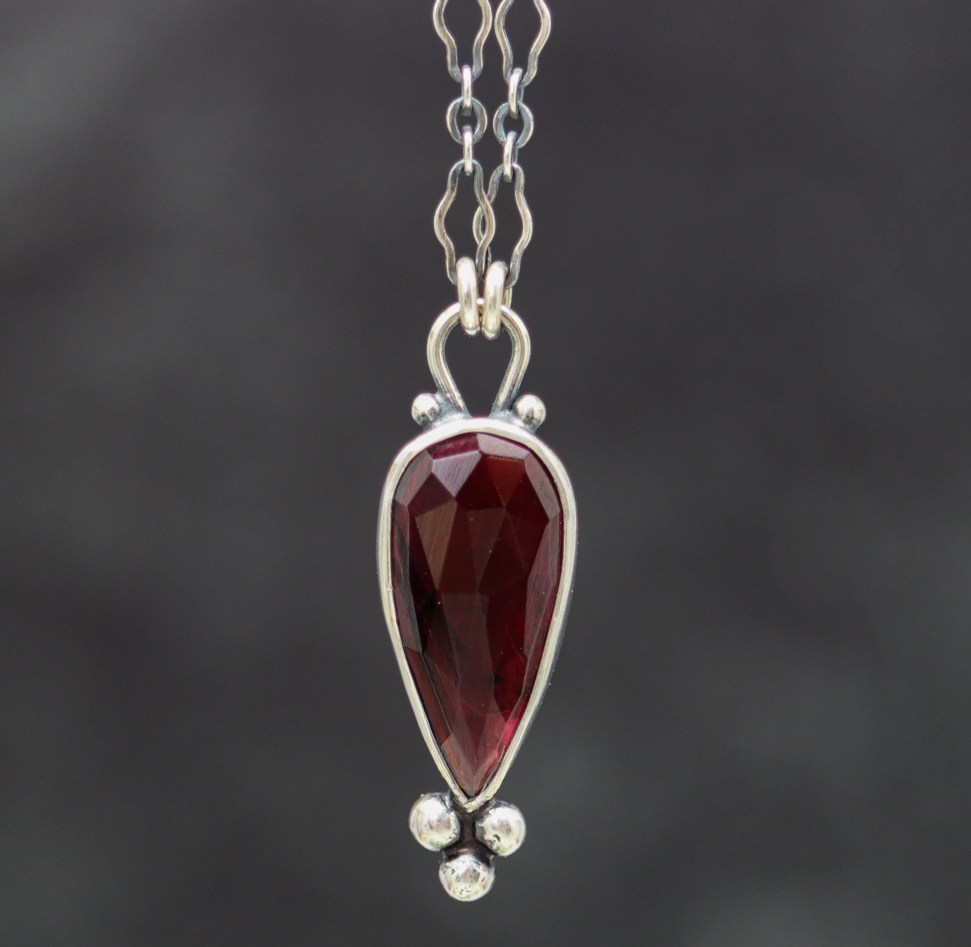 A sterling silver pendant necklace with a garnet centerpiece and small silver ball accents, featuring a textured backplate.