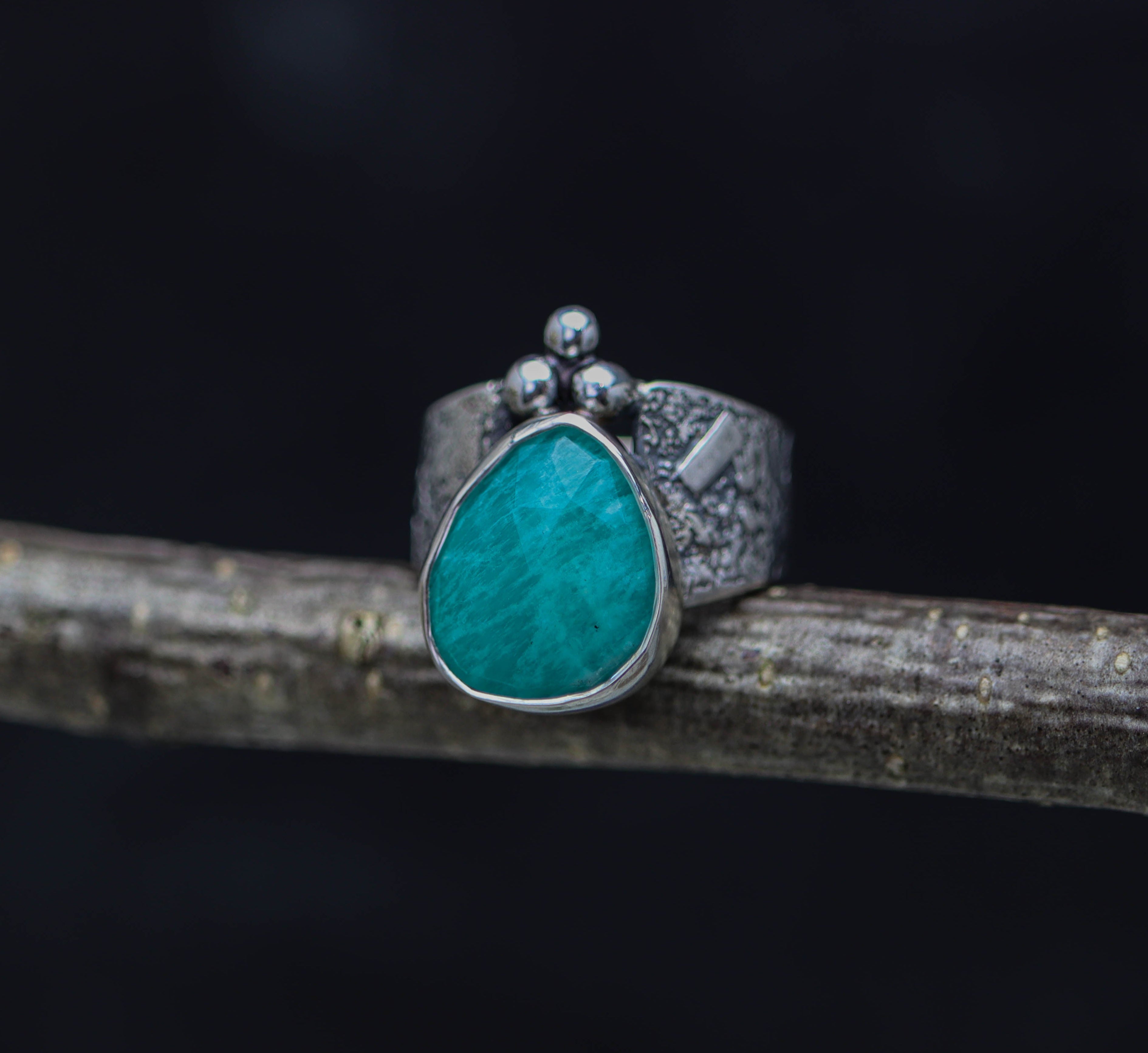 A wide band sterling silver ring with a textured surface and a rose cut amazonite gemstone set on top, displayed on a wooden branch against a black background.