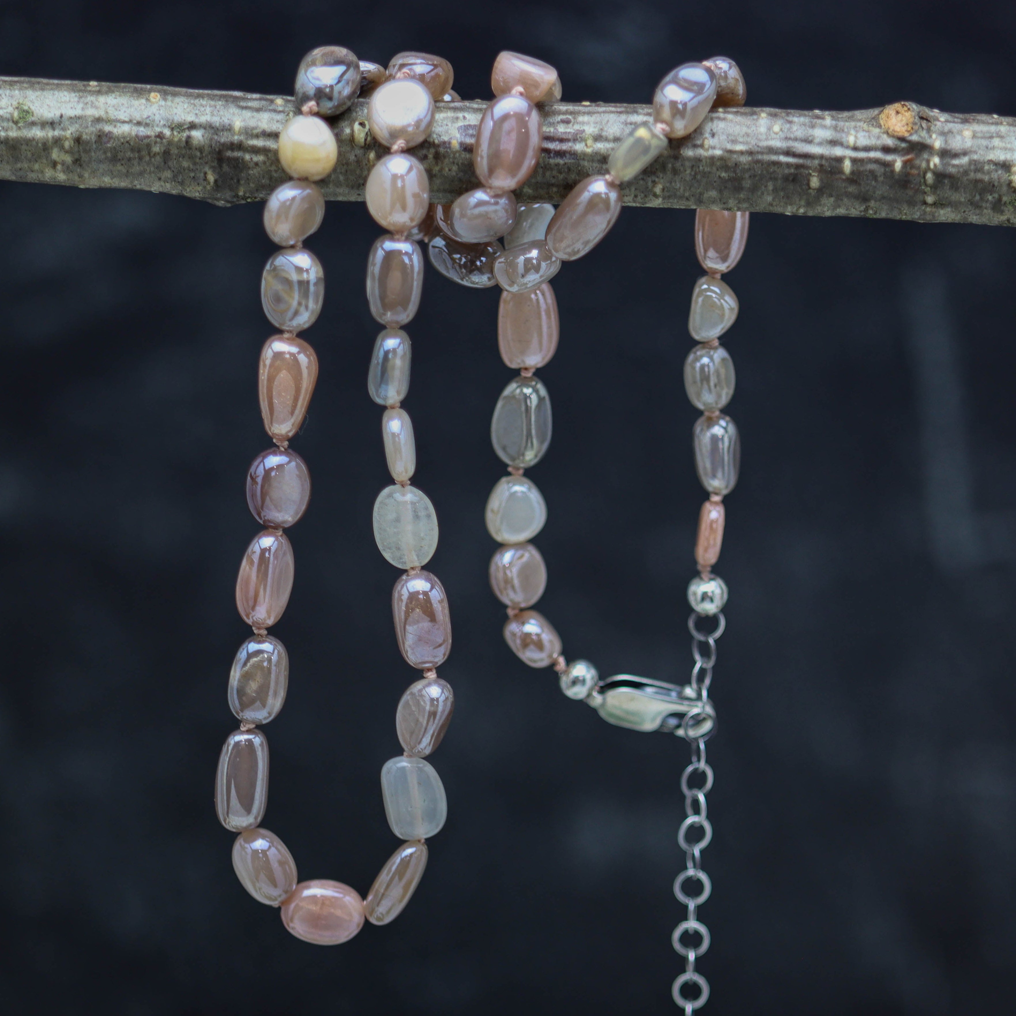 A hand knotted bead necklace with sterling silver closure, featuring silvery flash moonstone beads in pinkish peach to grey to almost white shades, displayed on a dark background.