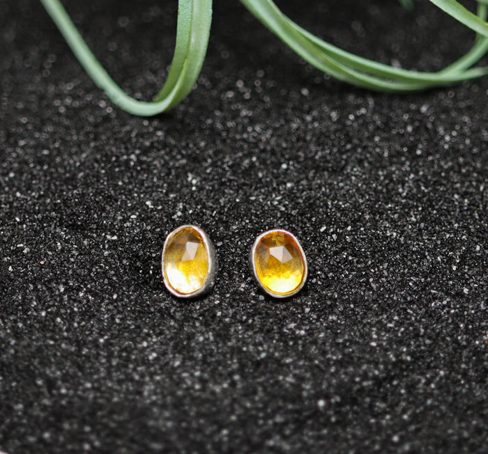 Pair of yellow citrine stud earrings on a black glittery surface with greenery.