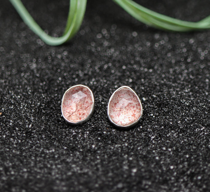 Pair of silver earrings with pinkish-red speckles on a dark textured surface.