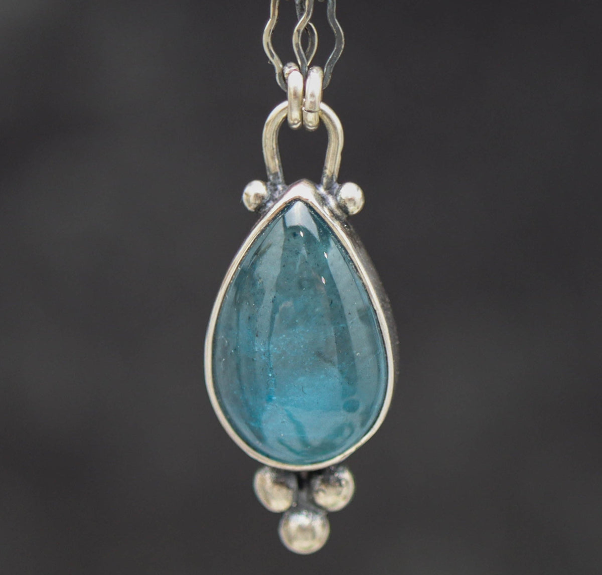 Teardrop-shaped blue topaz gemstone pendant on a chain against a dark background