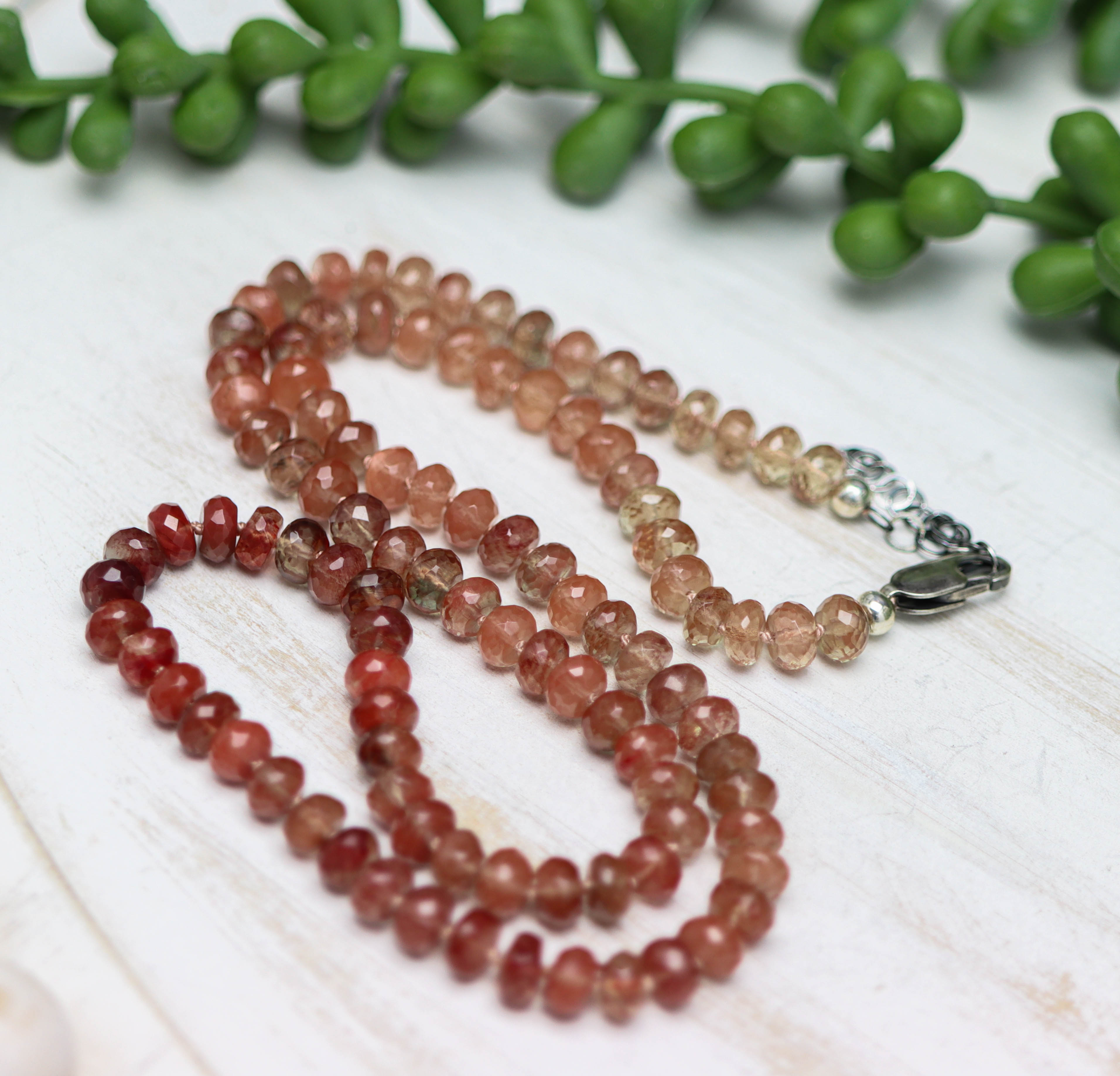 Beaded necklace with multicolored stones on a light wooden surface with green leaves in the background