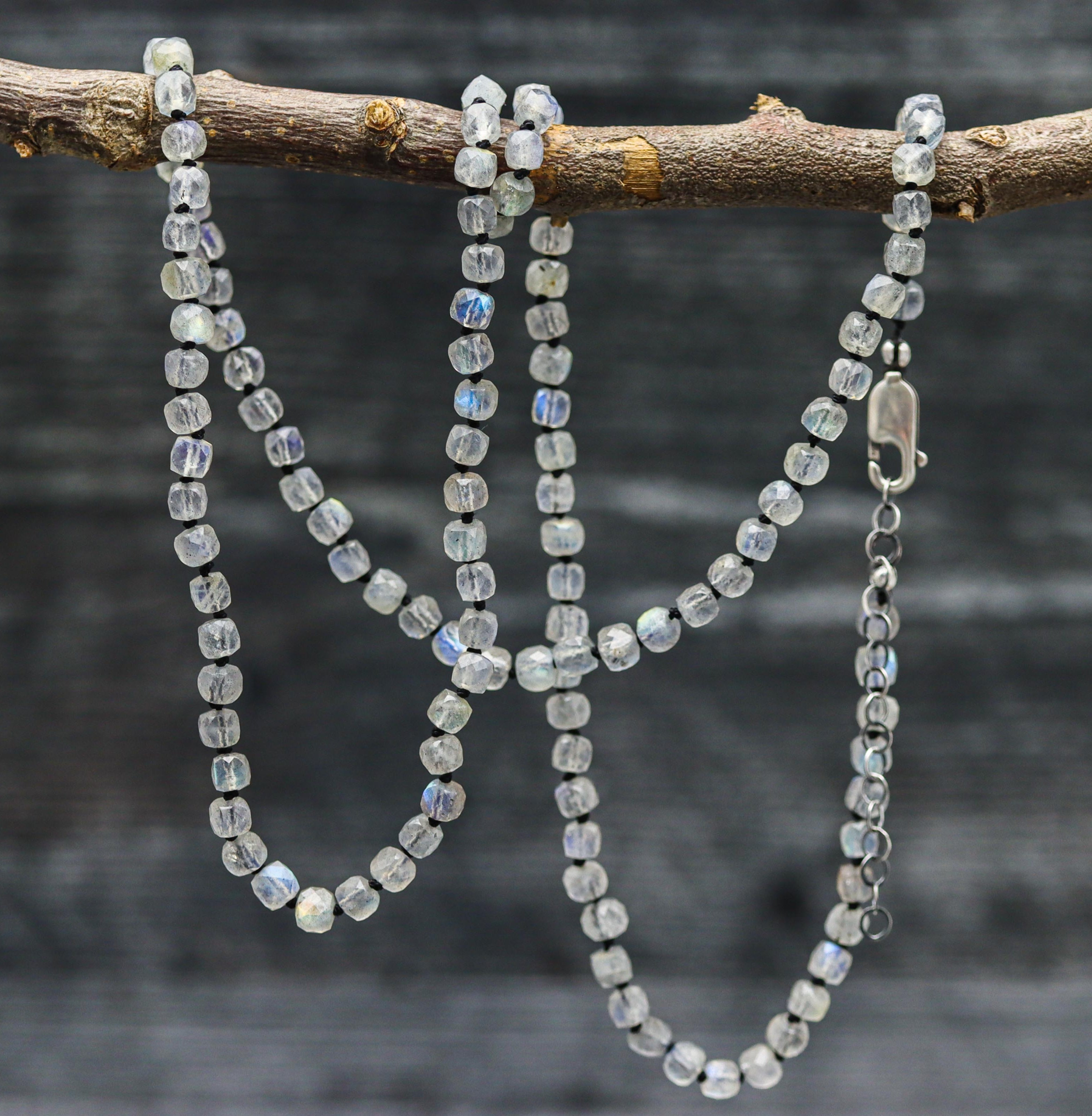 A hand knotted sterling silver necklace featuring faceted cube-shaped labradorite stones, displayed on a branch against a wooden backdrop.