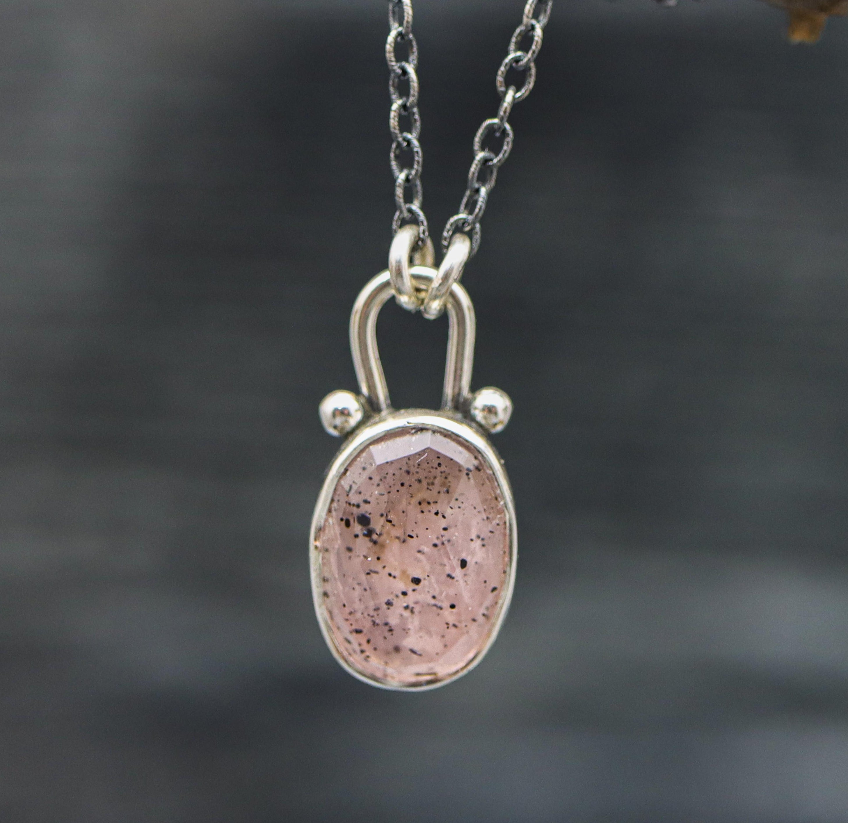 A sterling silver pendant necklace with a rose quartz gemstone and small silver ball accents, displayed against an outdoor background.